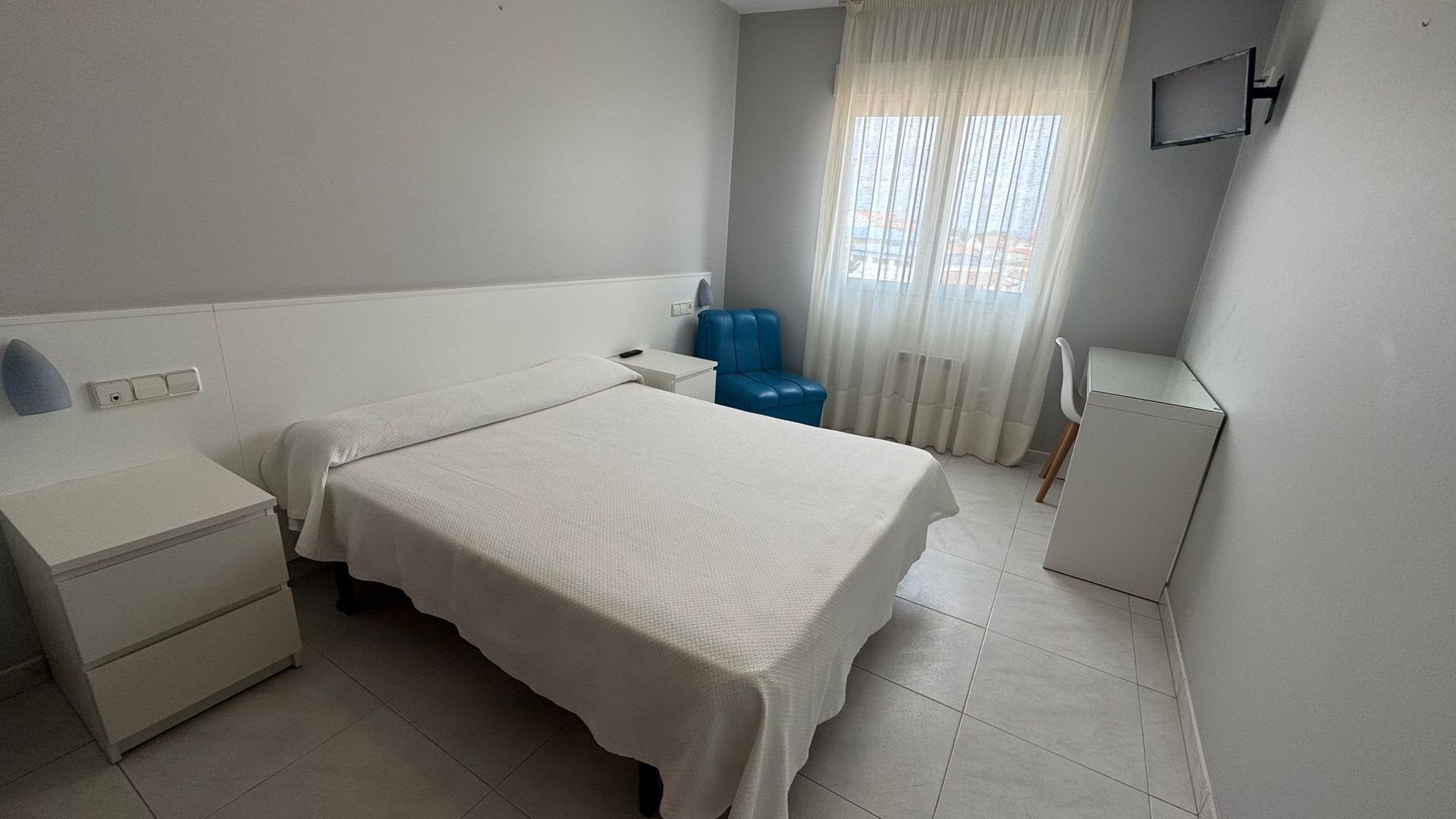 A modern, brightly lit hotel room with a neatly made white double bed, a white nightstand, a blue chair, and a desk.