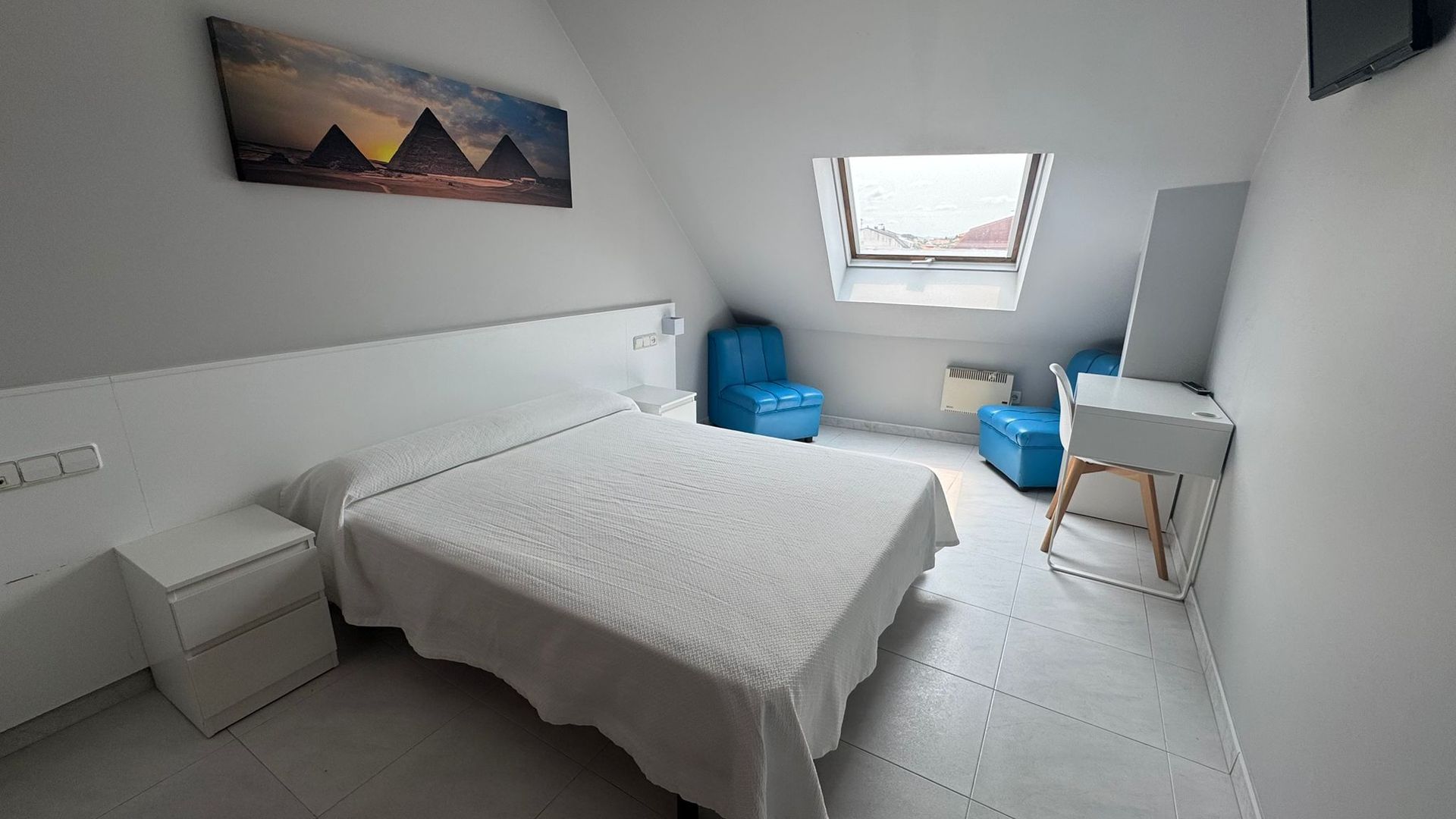 A minimalist bedroom with a double bed, white furniture, a pyramid wall art, and blue accent chairs under a skylight.