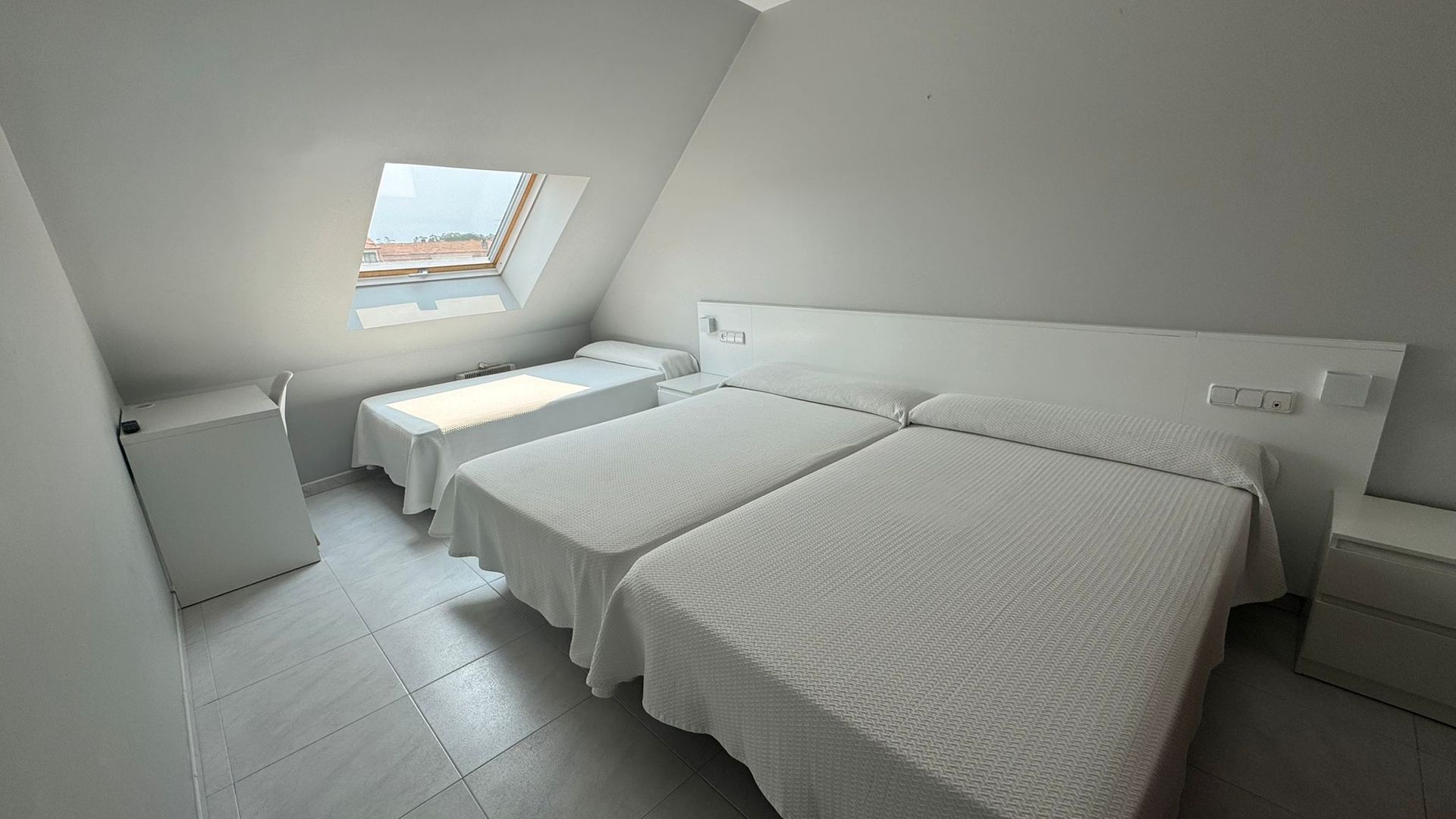 A minimalist hotel room with three white beds, white walls, light tile floors, and a slanted roof with a skylight window.