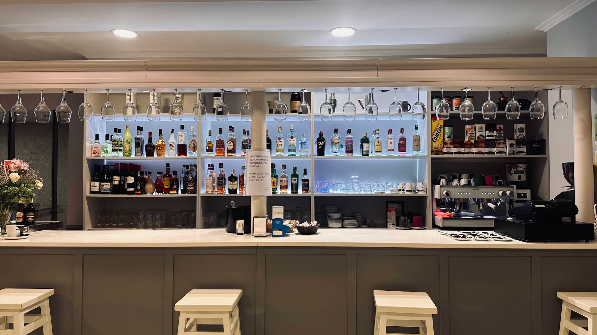 A modern, well-lit bar with shelves of various bottles, glassware hanging above, and four white stools at the counter.