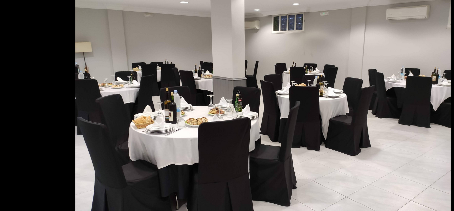A bar area with shelves of bottles, white bar stools, and round tables with blue-covered chairs in a neutral-toned room.