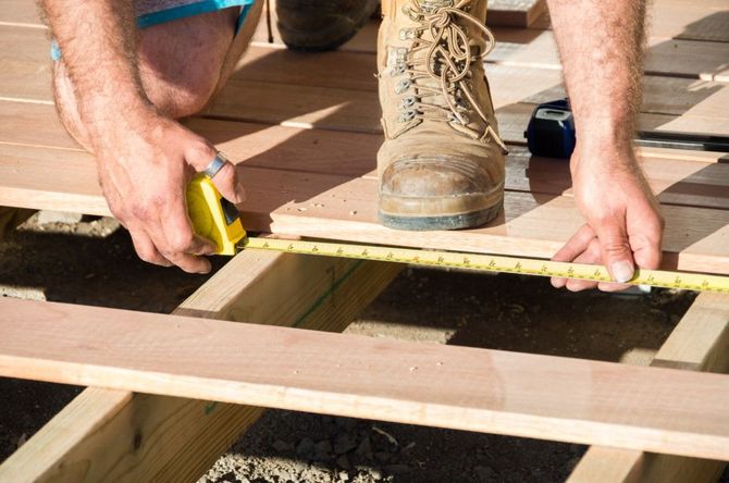 Person kneeling, measuring wooden deck boards with a yellow tape measure, outdoors.