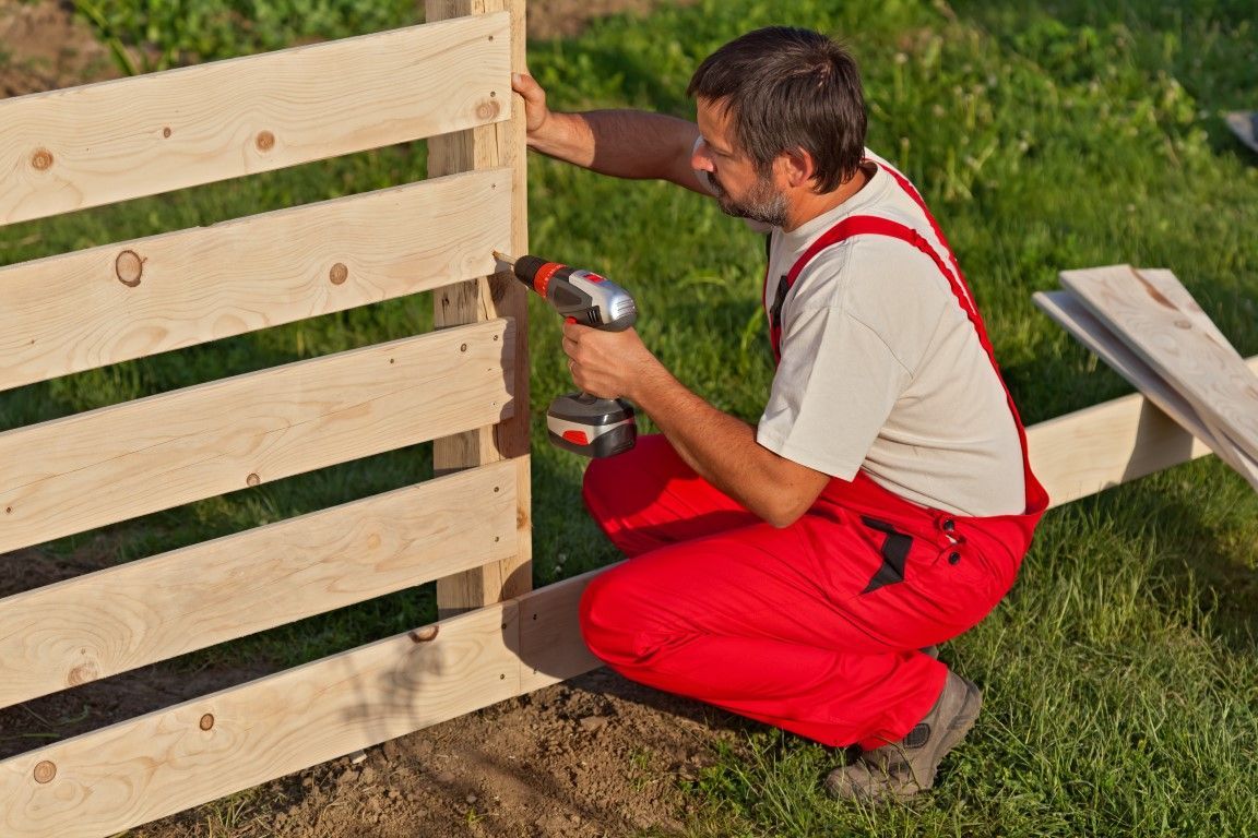 Man in red overalls using a power drill to assemble a wooden fence outdoors.