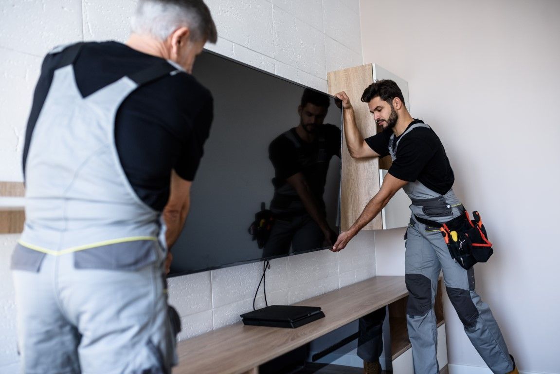 Two workers installing a large TV on a wall above a wooden console.
