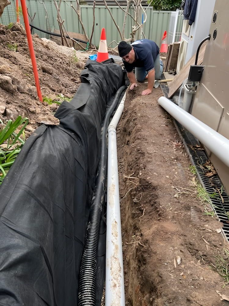 A Man is Kneeling Down in the Dirt Next to a Pipe — Around Town Plumbing Services in Canberra, ACT