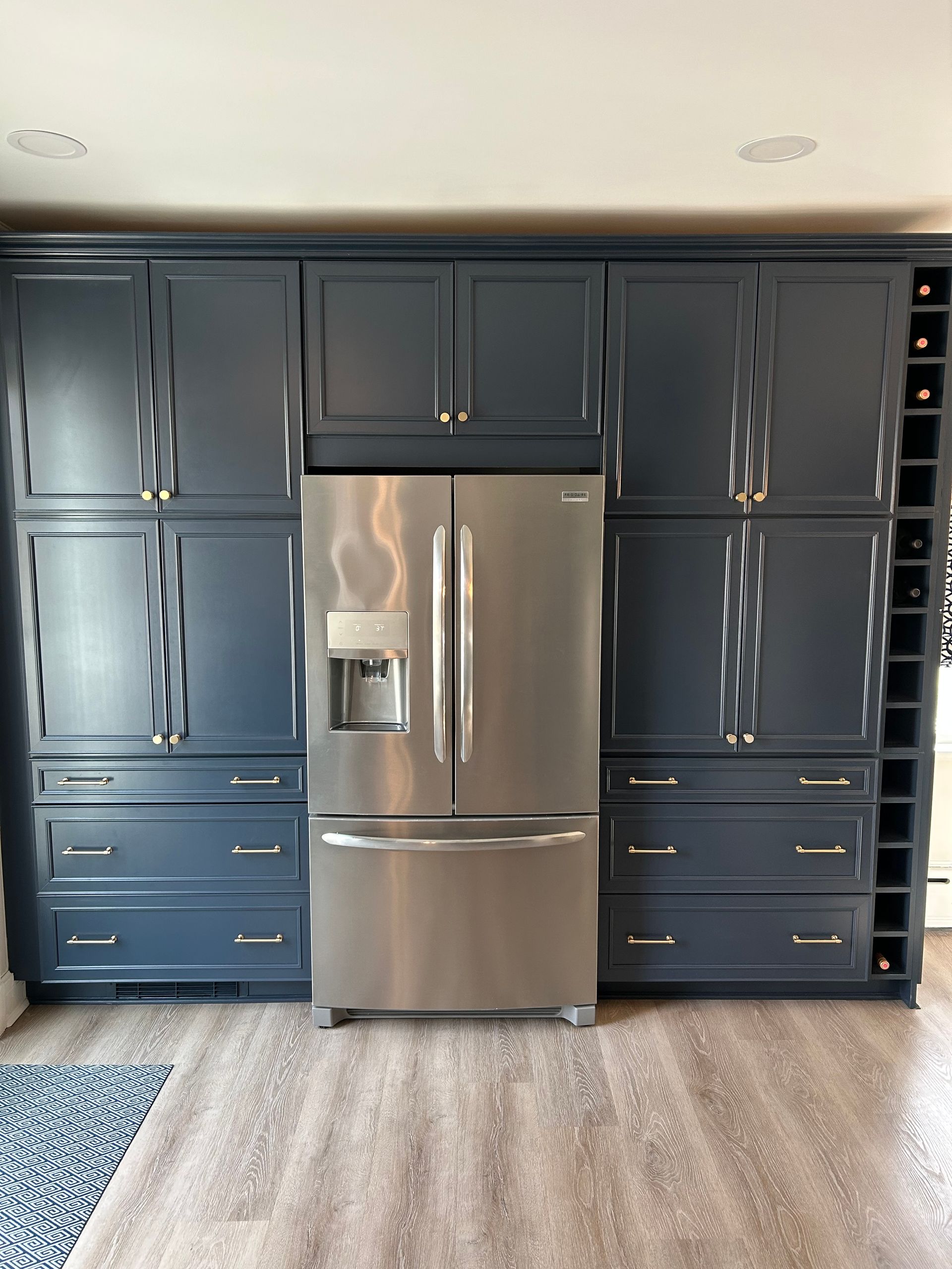 Blue kitchen cabinets surround a stainless steel refrigerator. A wine rack is on the right.