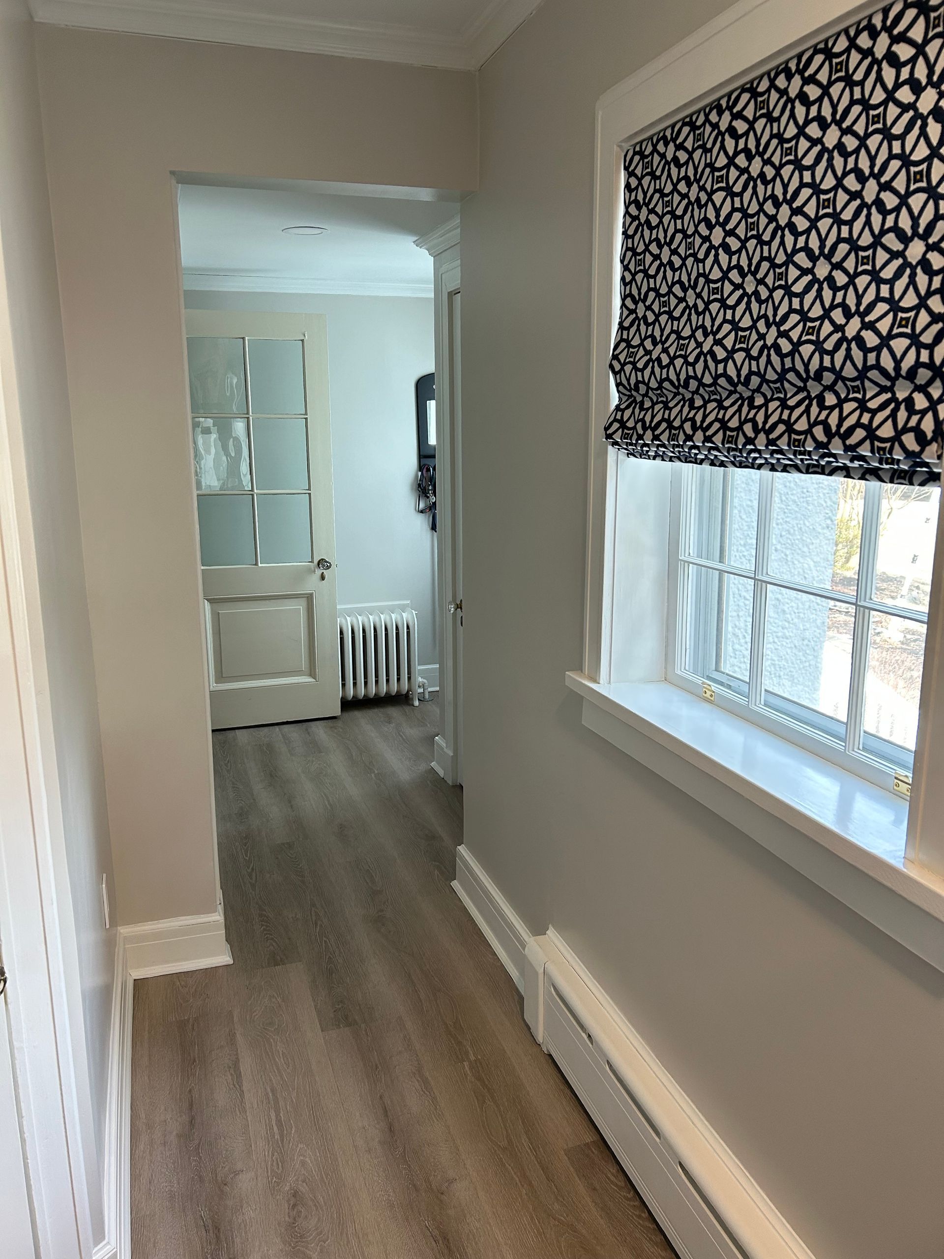 Narrow hallway with pale walls, light-colored flooring, and a window with patterned shade.