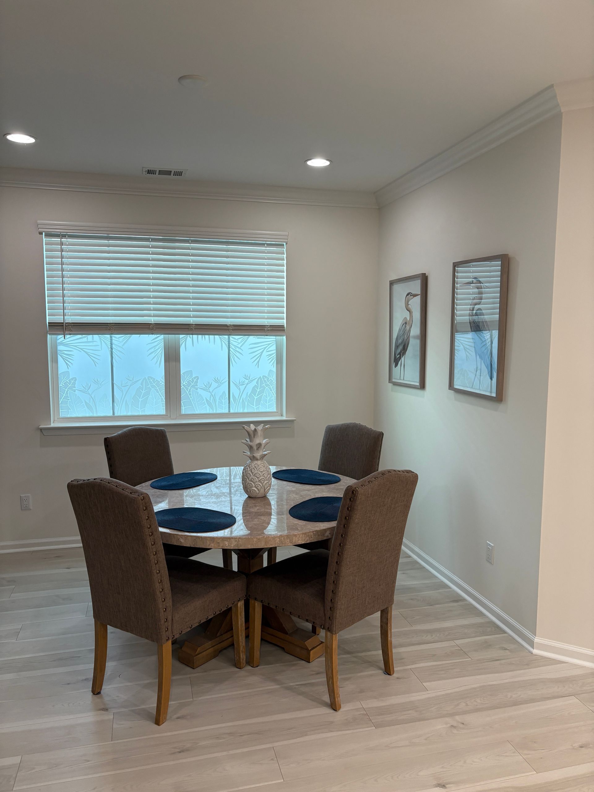 Dining room with round table, four chairs, and framed art. Window with blinds provides natural light.
