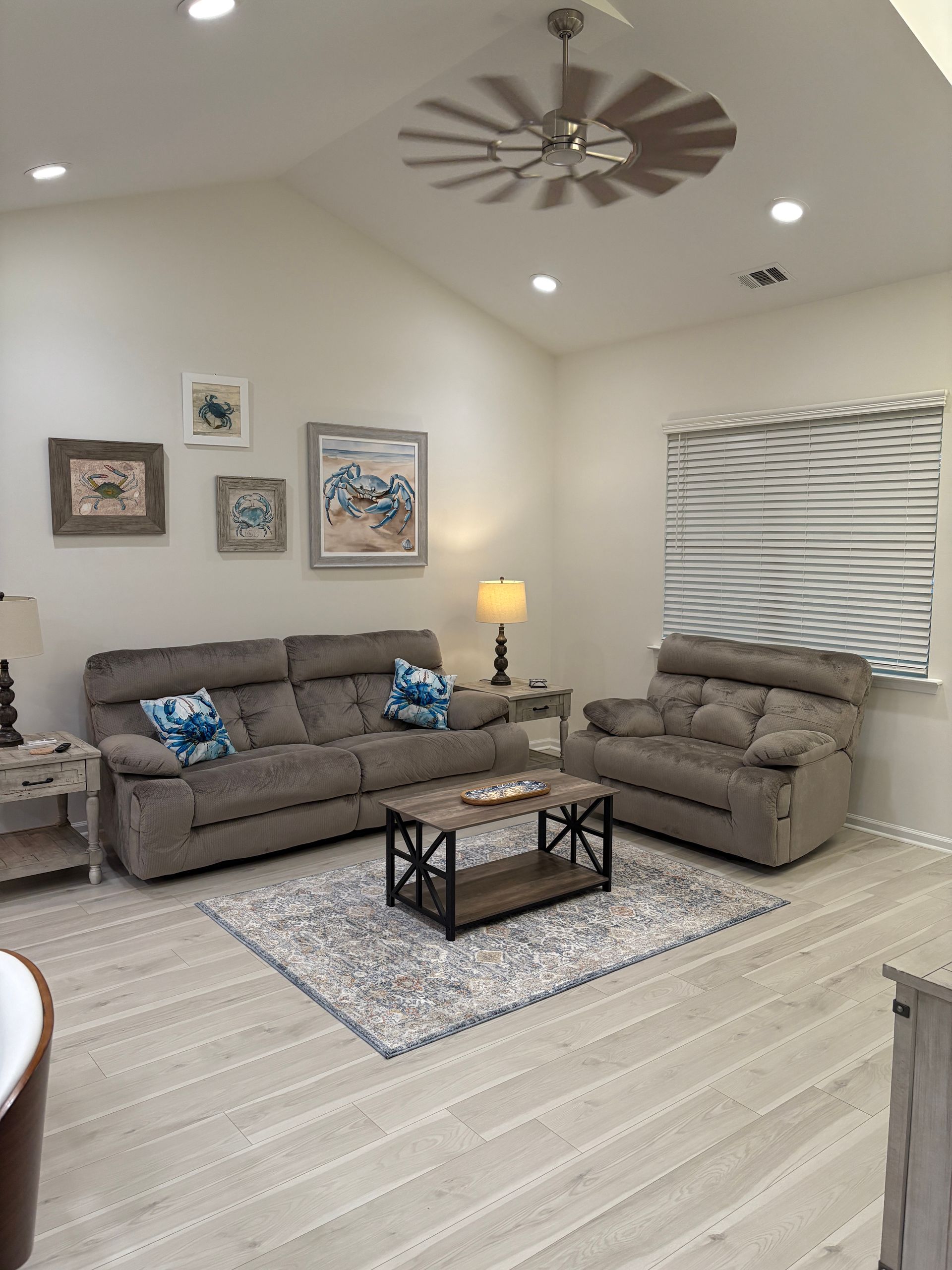 Living room with gray sofa, chair, and rug, light walls, and artwork. Ceiling fan and window.