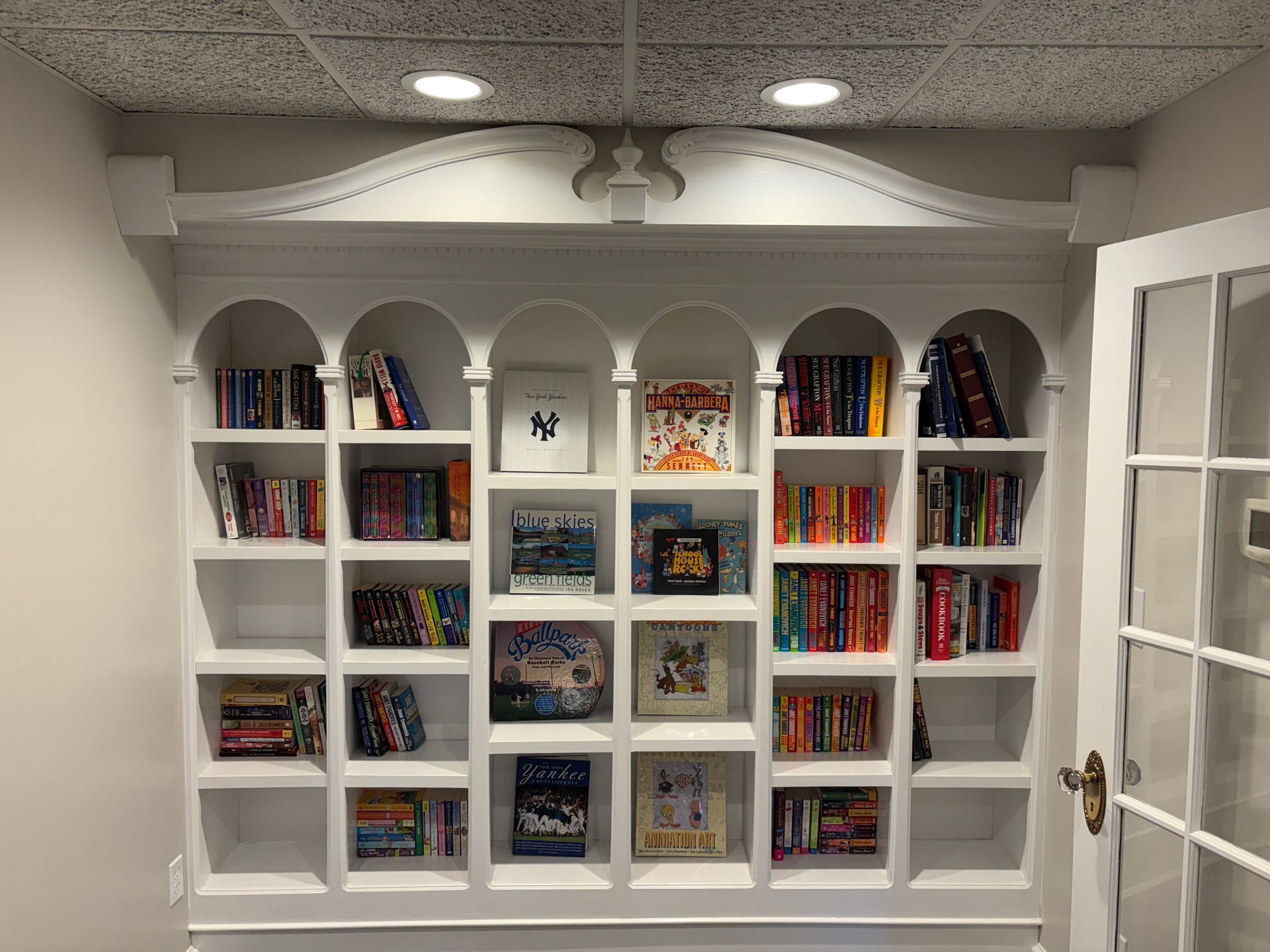 White built-in bookshelf filled with books, arched top, and decorative molding. Next to a white door.