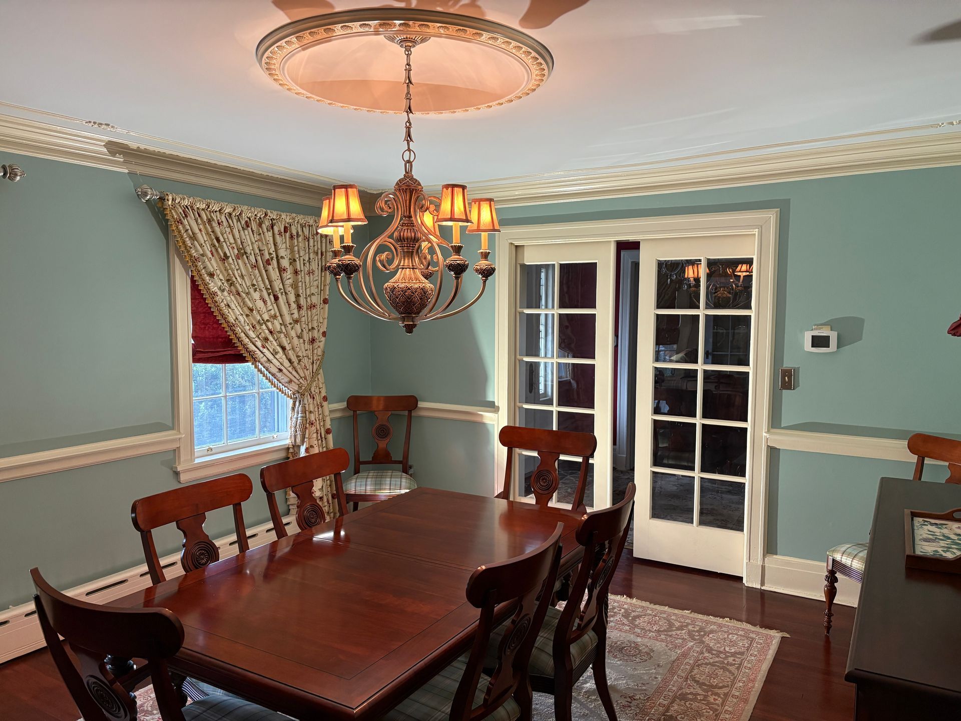 Dining room with wooden table, chairs, chandelier, blue walls, and window with curtains.