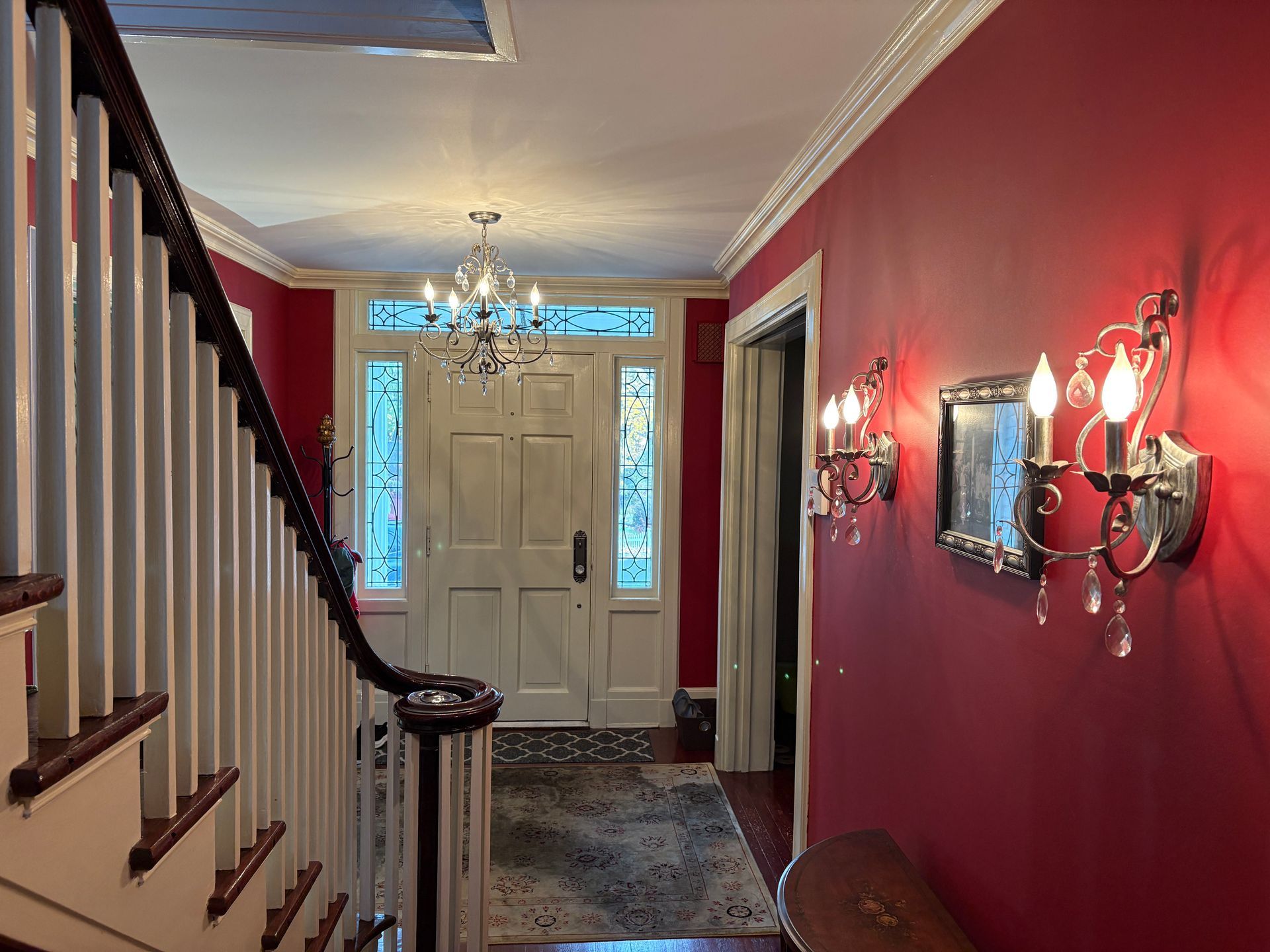 Interior hallway with red walls, staircase, chandelier, and sconces. Door at end with sidelights.