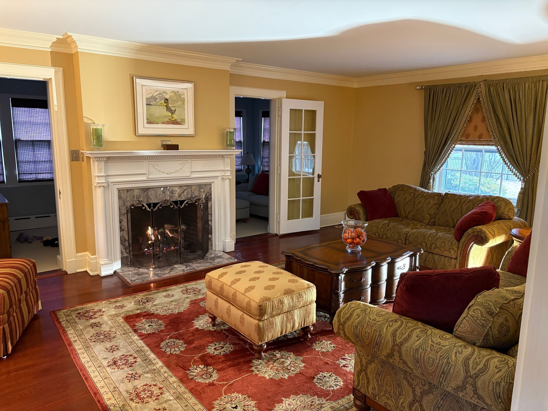 Cozy living room with fireplace, couches, red rug, and ottoman. Golden walls and ornate trim.