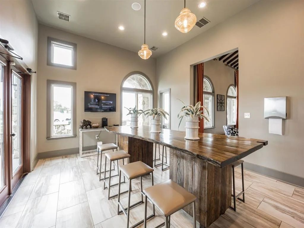 Lounge area with a long wooden table, bar stools, and vases. Windows and a TV are visible.