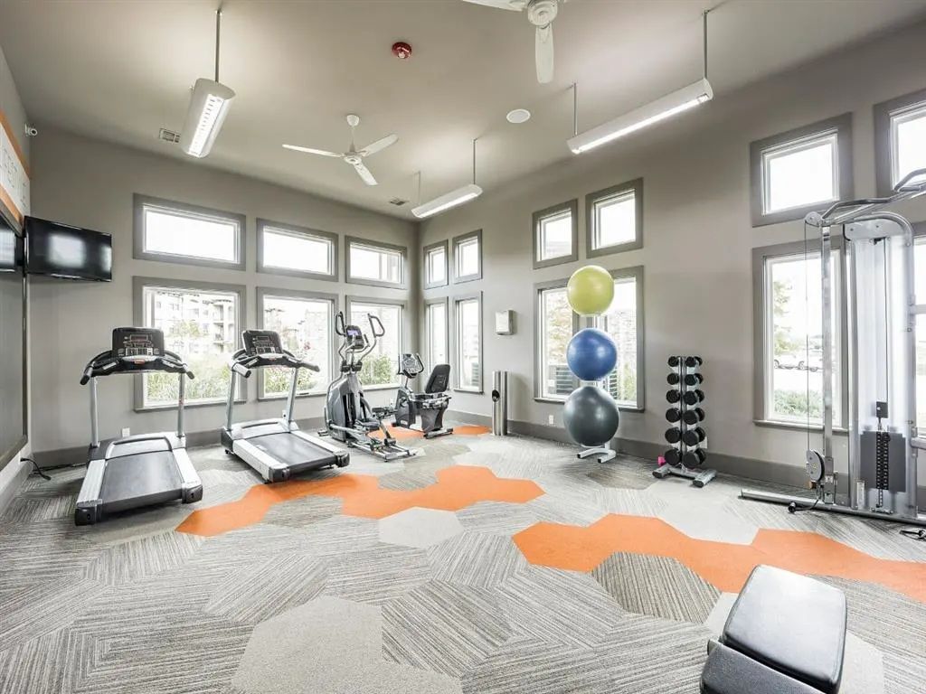 Gym interior with exercise equipment, including treadmills, weights, and a weight machine. Windows and overhead lights illuminate the space.