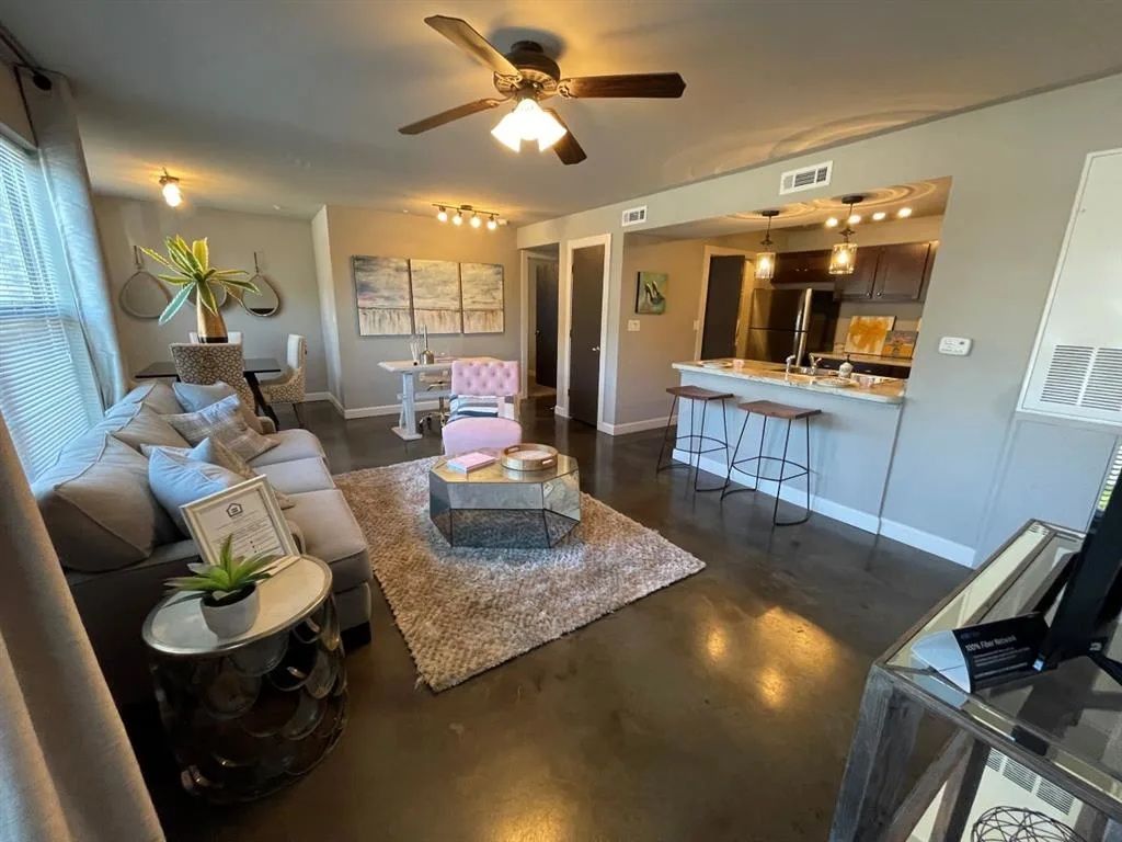 Living room with gray sofa, pink chair, and rug. Open kitchen with bar seating, and a ceiling fan.