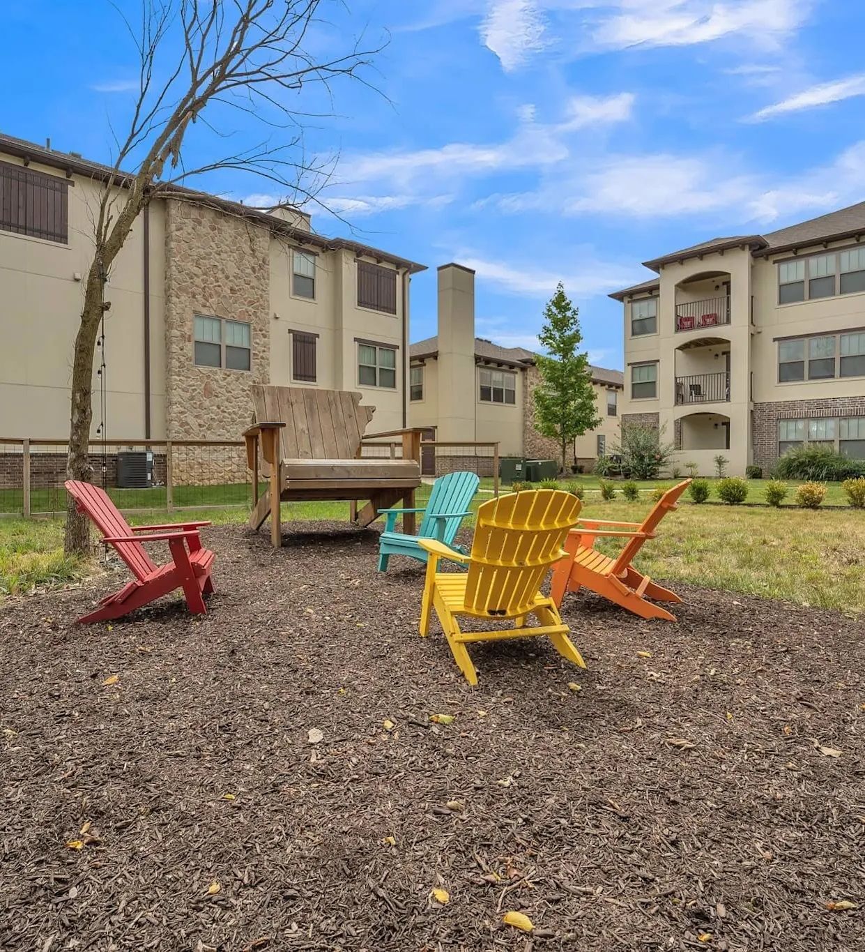Outdoor seating area with colorful Adirondack chairs in front of apartment buildings under a blue sky.