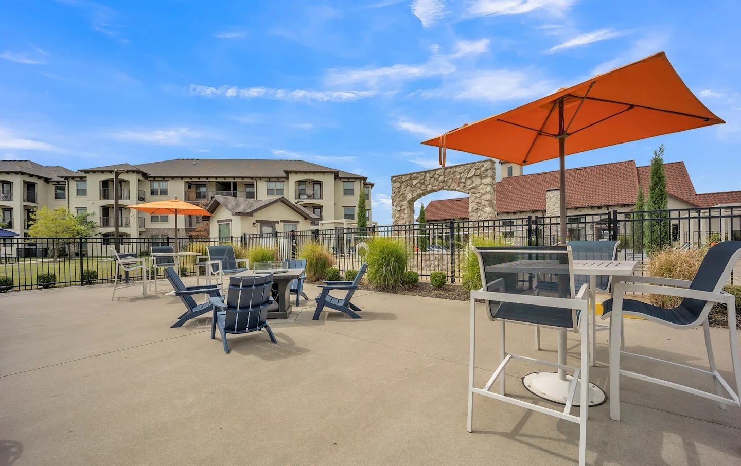 Outdoor grilling area with tables, chairs, and orange umbrellas near an apartment building under a blue sky.