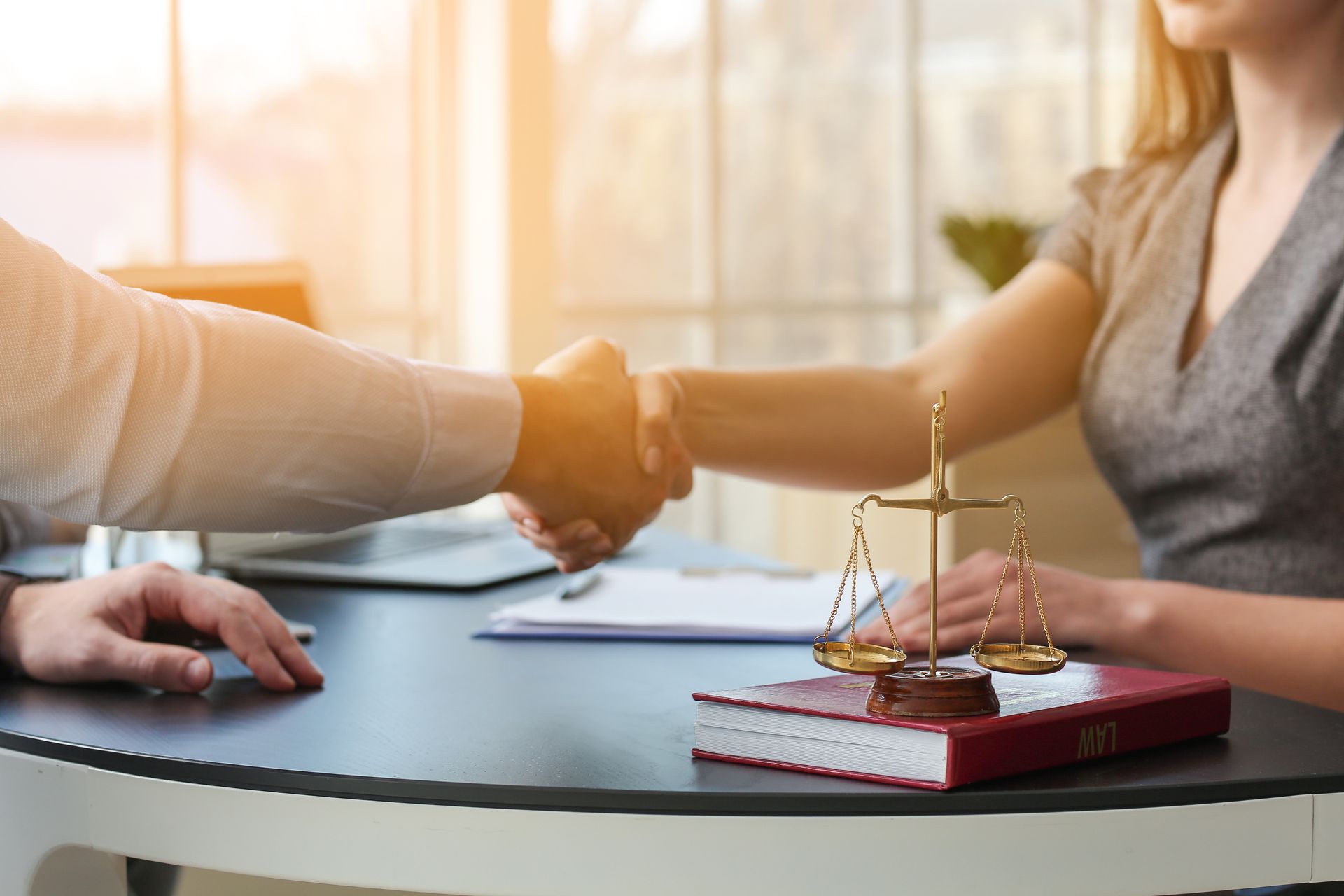 A lawyer and a client shake hands over a desk. A book and the scales of justice are on the desk.