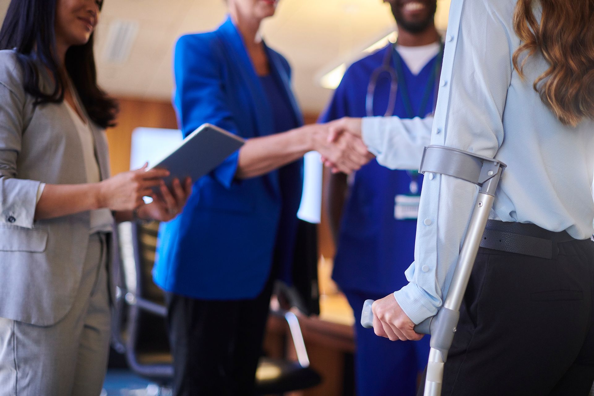 Person with crutch in meeting as two others shake hands and one holds a tablet.