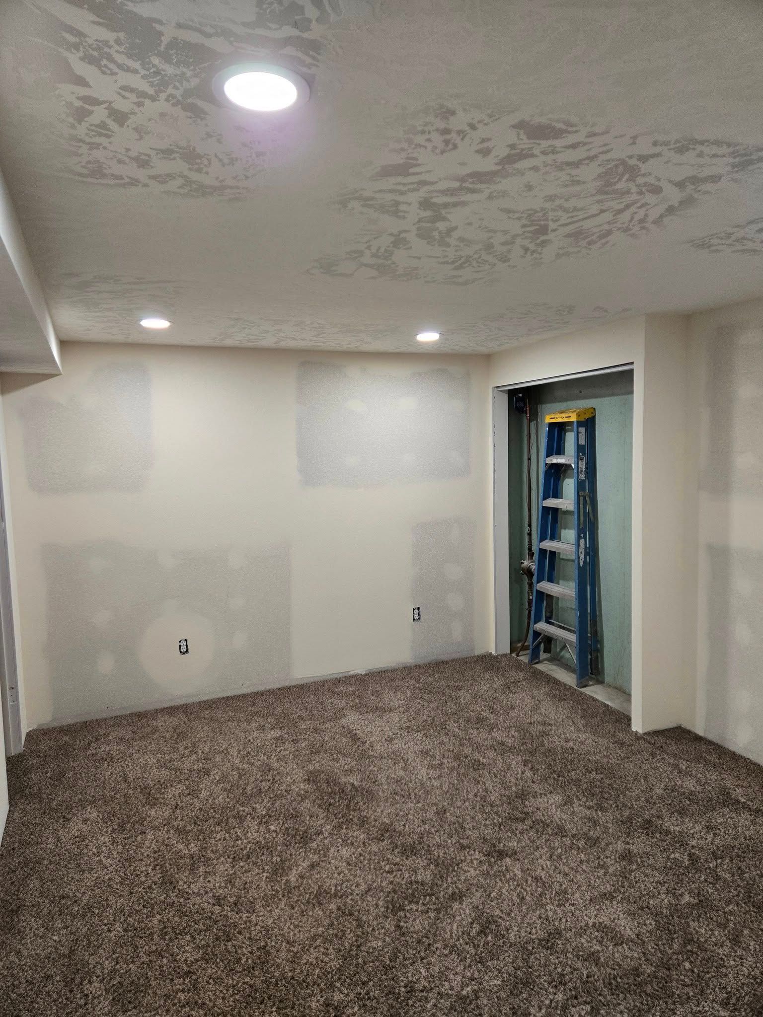 Room with brown carpet, white walls with drywall patches, recessed lights, and a blue ladder.