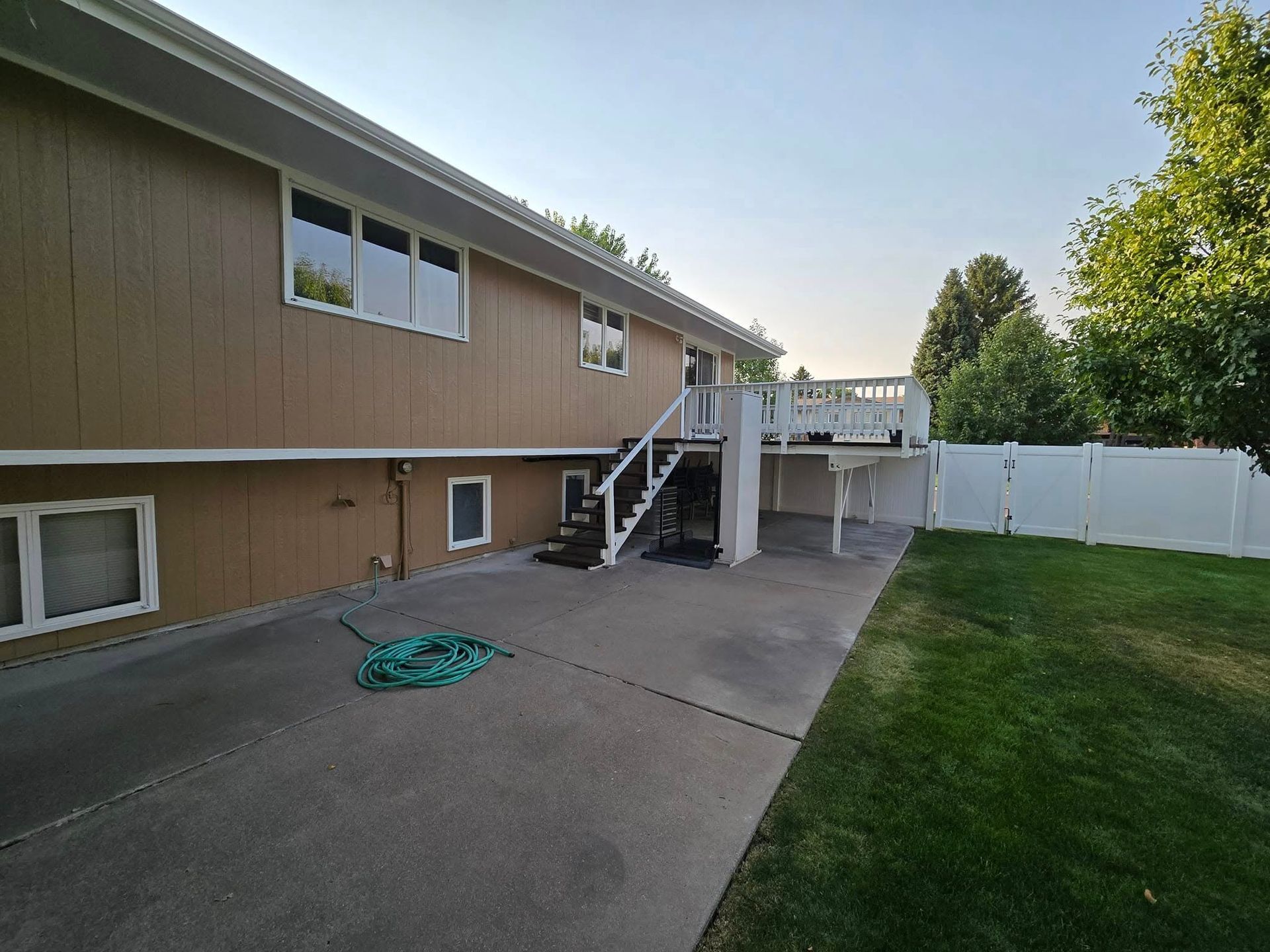 A two-story house with a deck and stairs, a concrete patio, and a green lawn with a hose.
