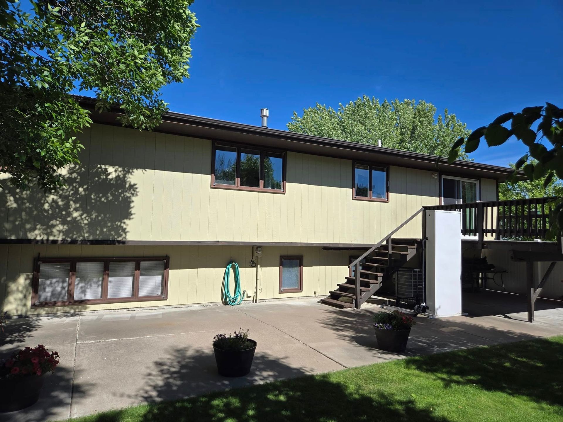 Two-story house with light yellow siding, brown trim, deck, and stairs. A garden hose and potted plants sit outside.
