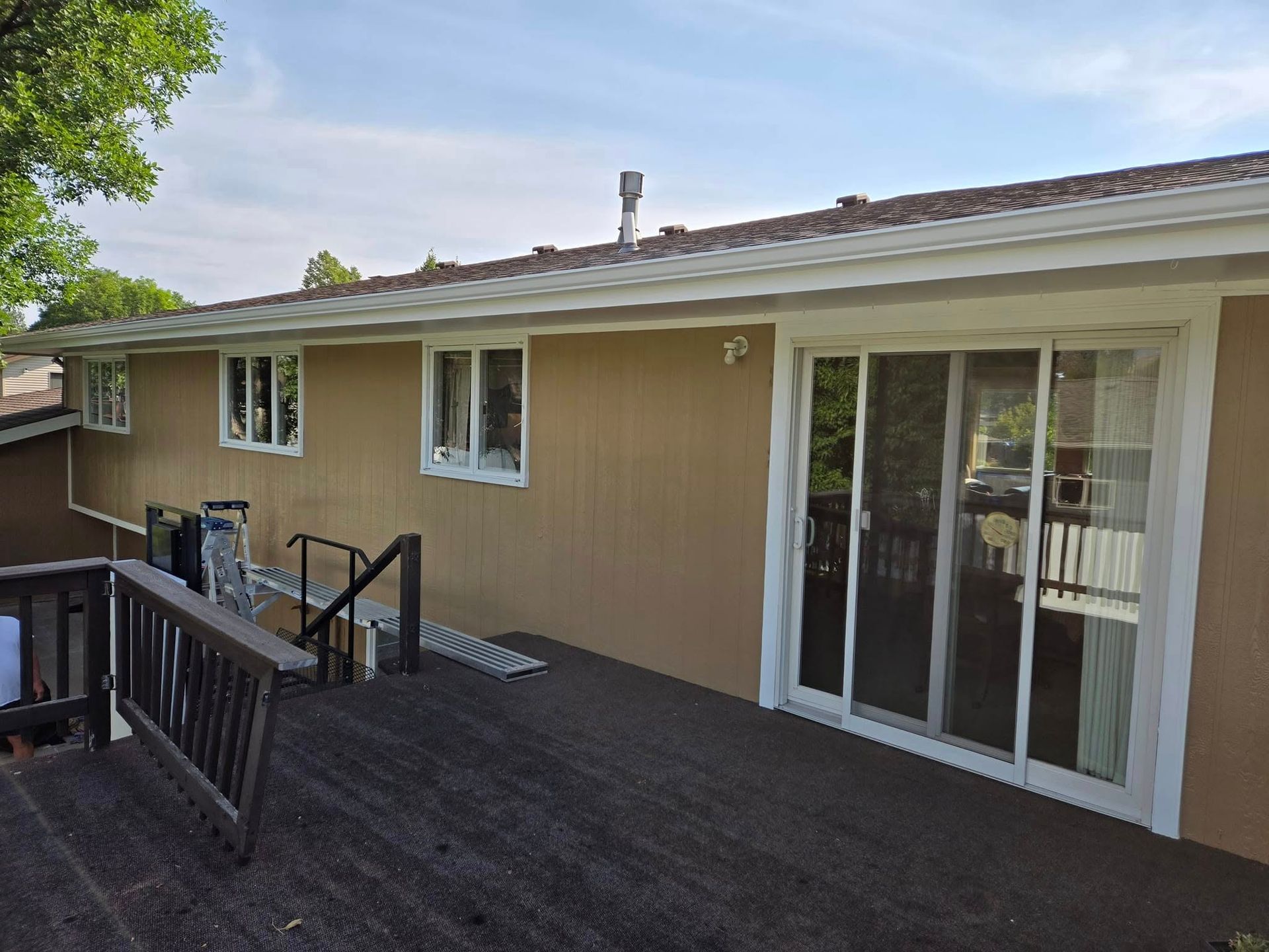 Brown house with deck, white-framed windows, and sliding glass door. Dark deck, blue sky.