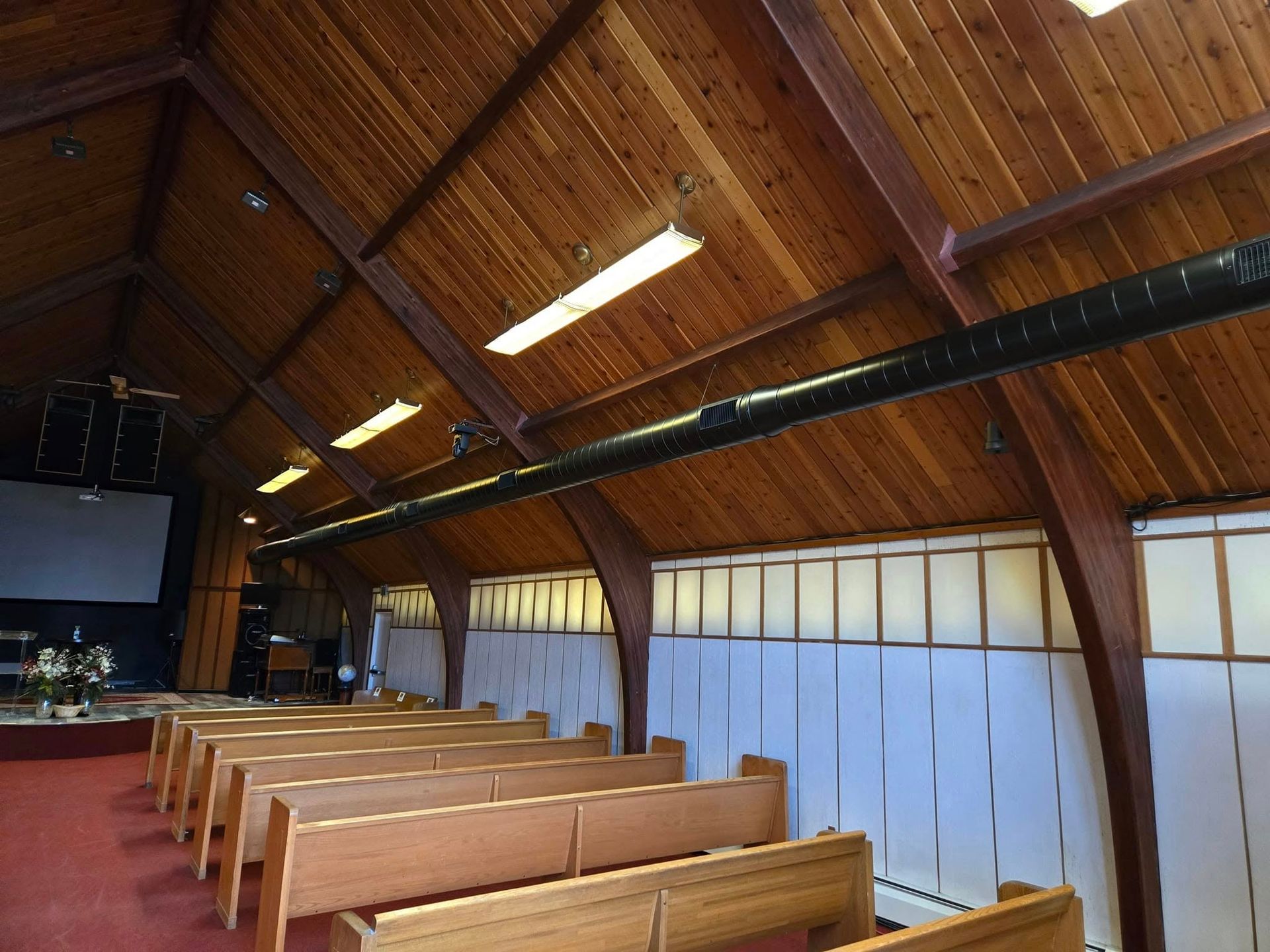Wooden church interior with rows of pews, arched ceiling, and stage with screen.