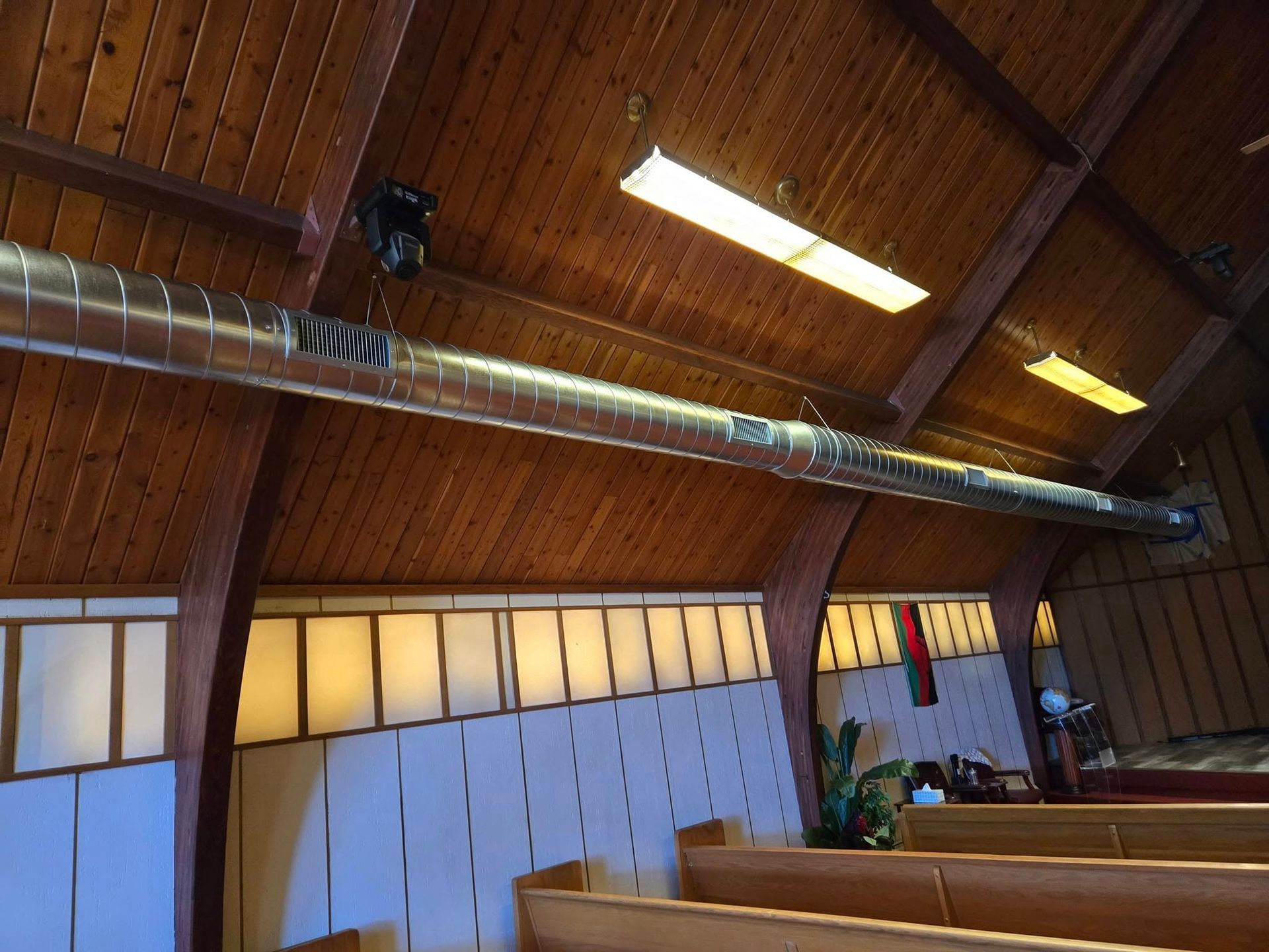 Wooden church ceiling with a metal air vent and two fluorescent lights.