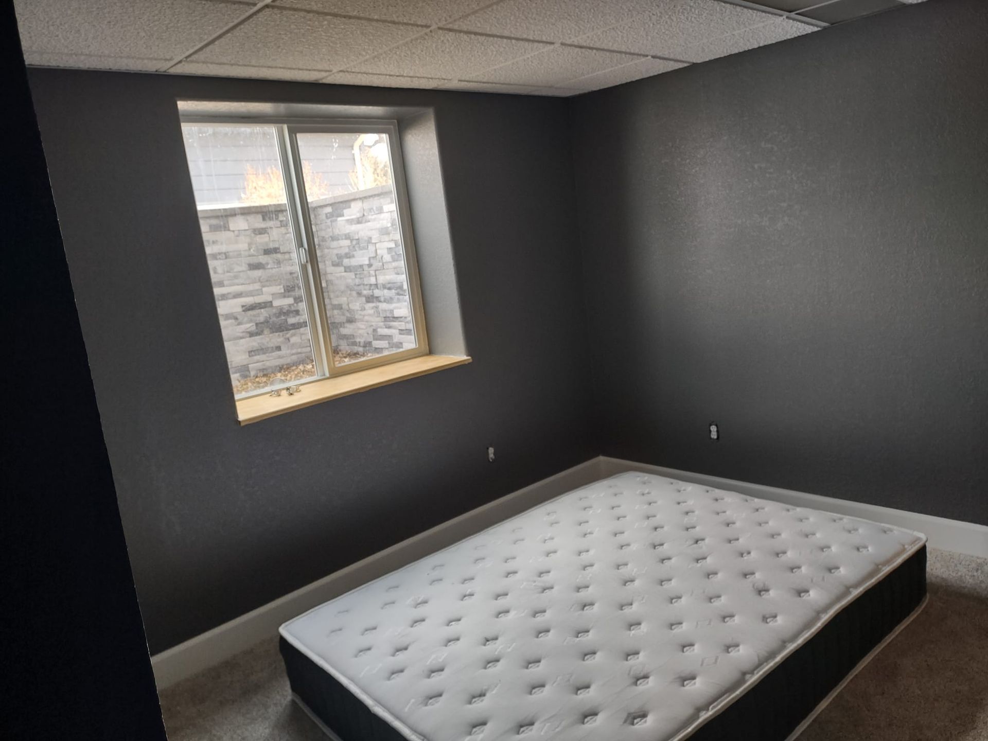 A dark gray bedroom with a mattress on the floor and a window with a stone-like wall outside.
