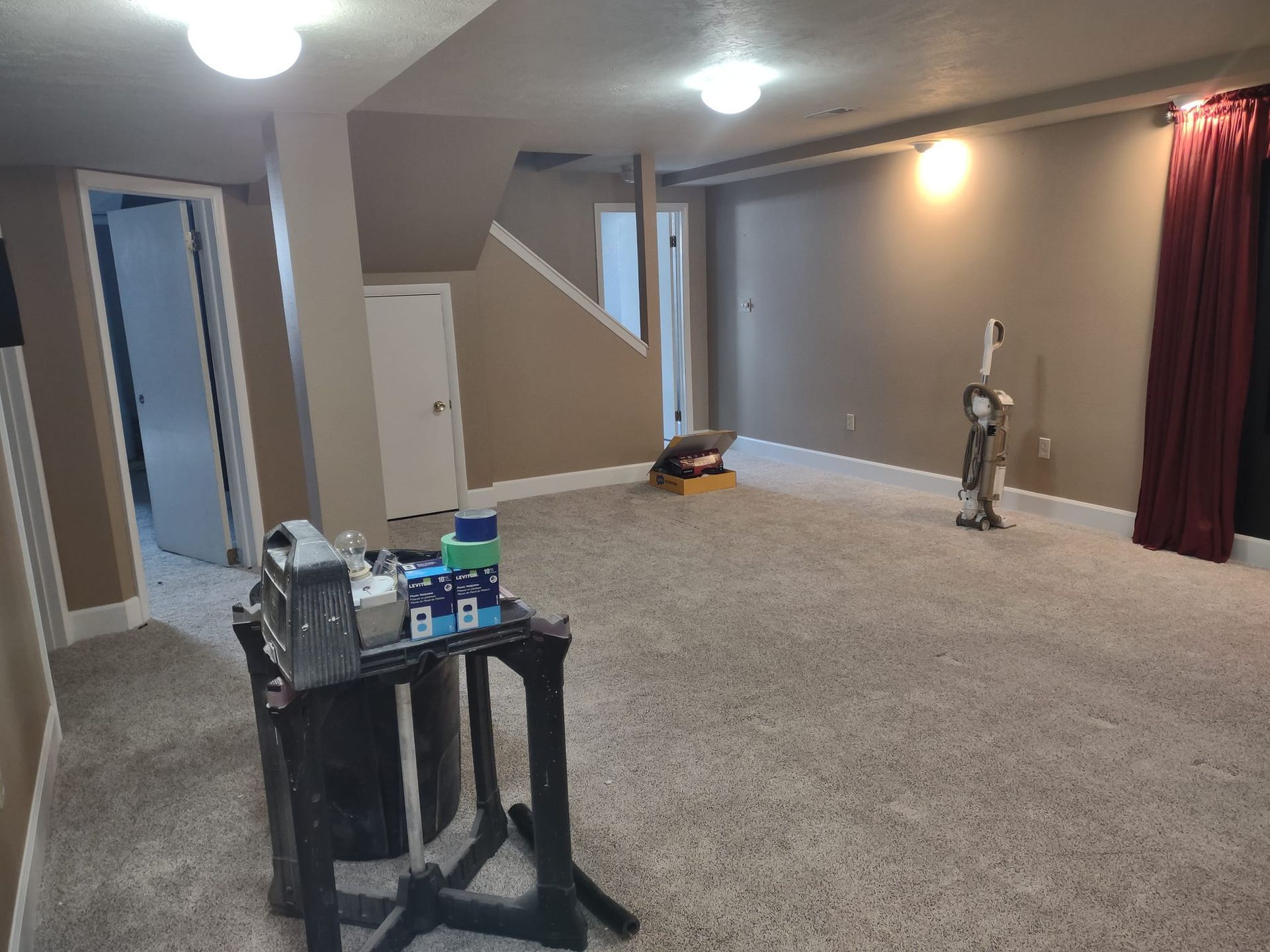 Basement room with carpet, tan walls, and a workstation; vacuum cleaner and curtains visible.