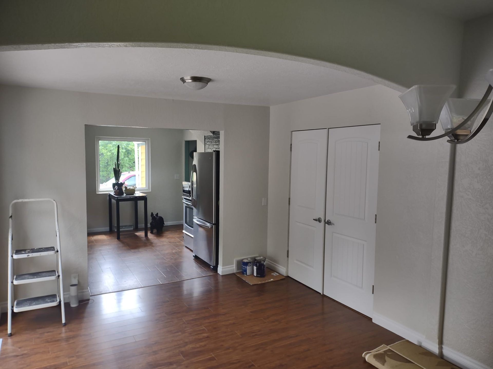 Interior view: Living room with open doorway to kitchen. Dark wood floor, white walls, ladder, and door.