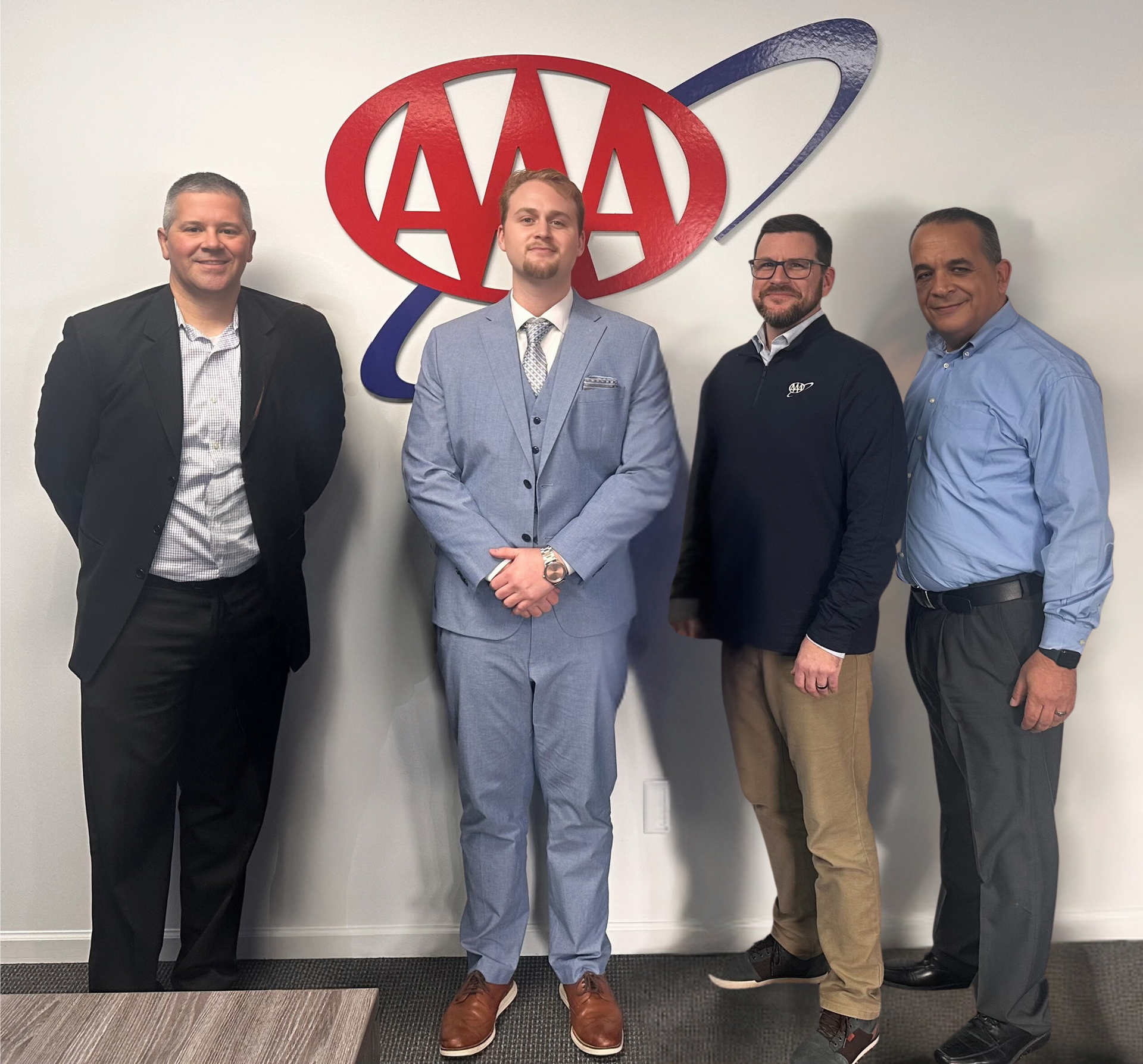 Four men pose in front of a AAA logo. One in a blue suit, others in business casual attire.