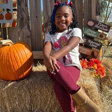 A little girl is sitting on a bale of hay next to a pumpkin.