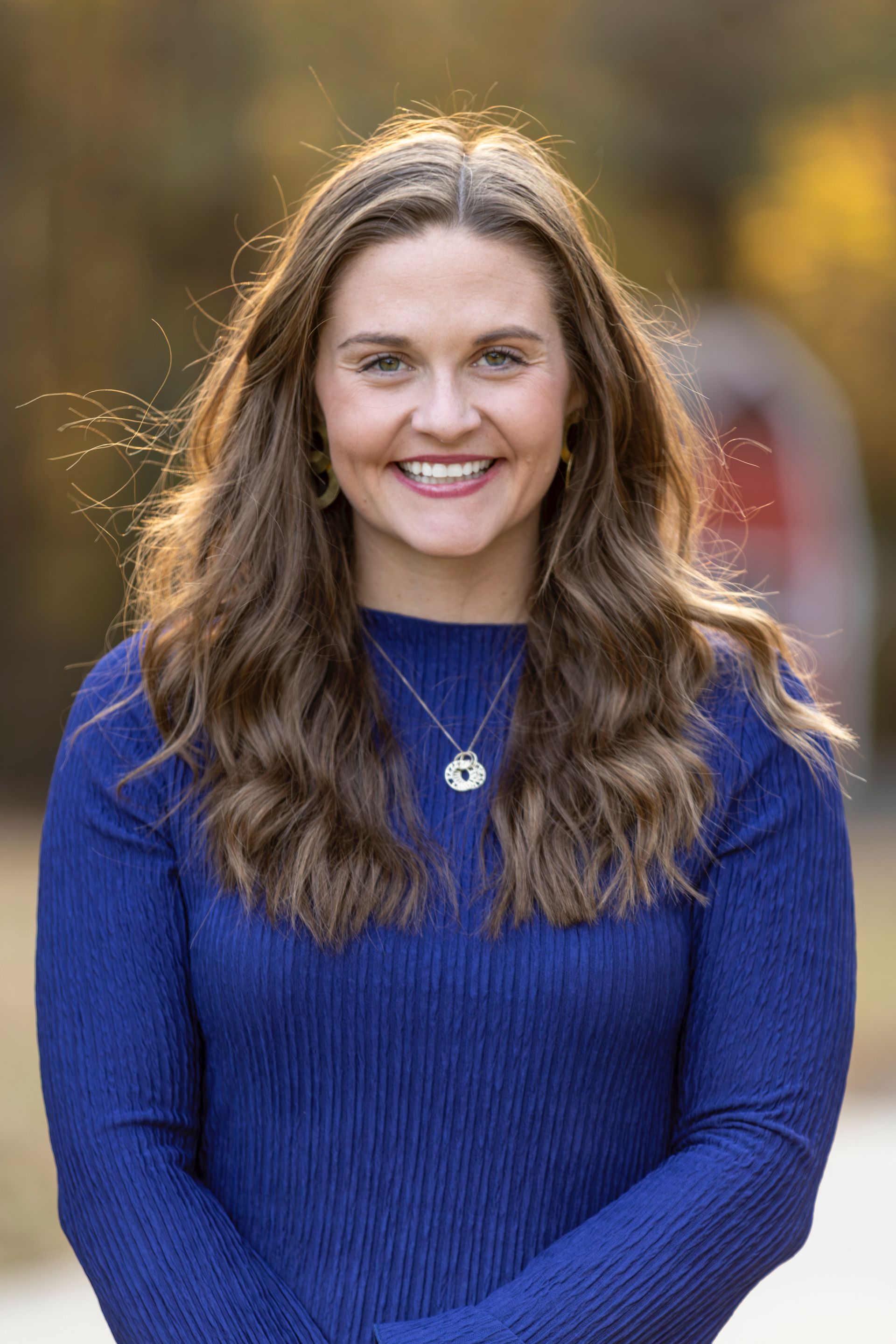 Woman with long brown hair smiling in blue sweater, wearing necklace, outside in front of blurred background.