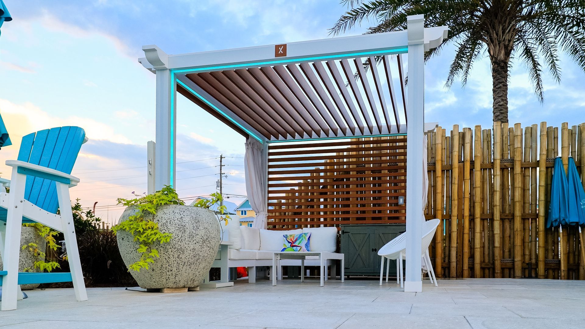 Pergola with seating area on a deck, light blue chairs, palm tree, bamboo fence, and setting sun.