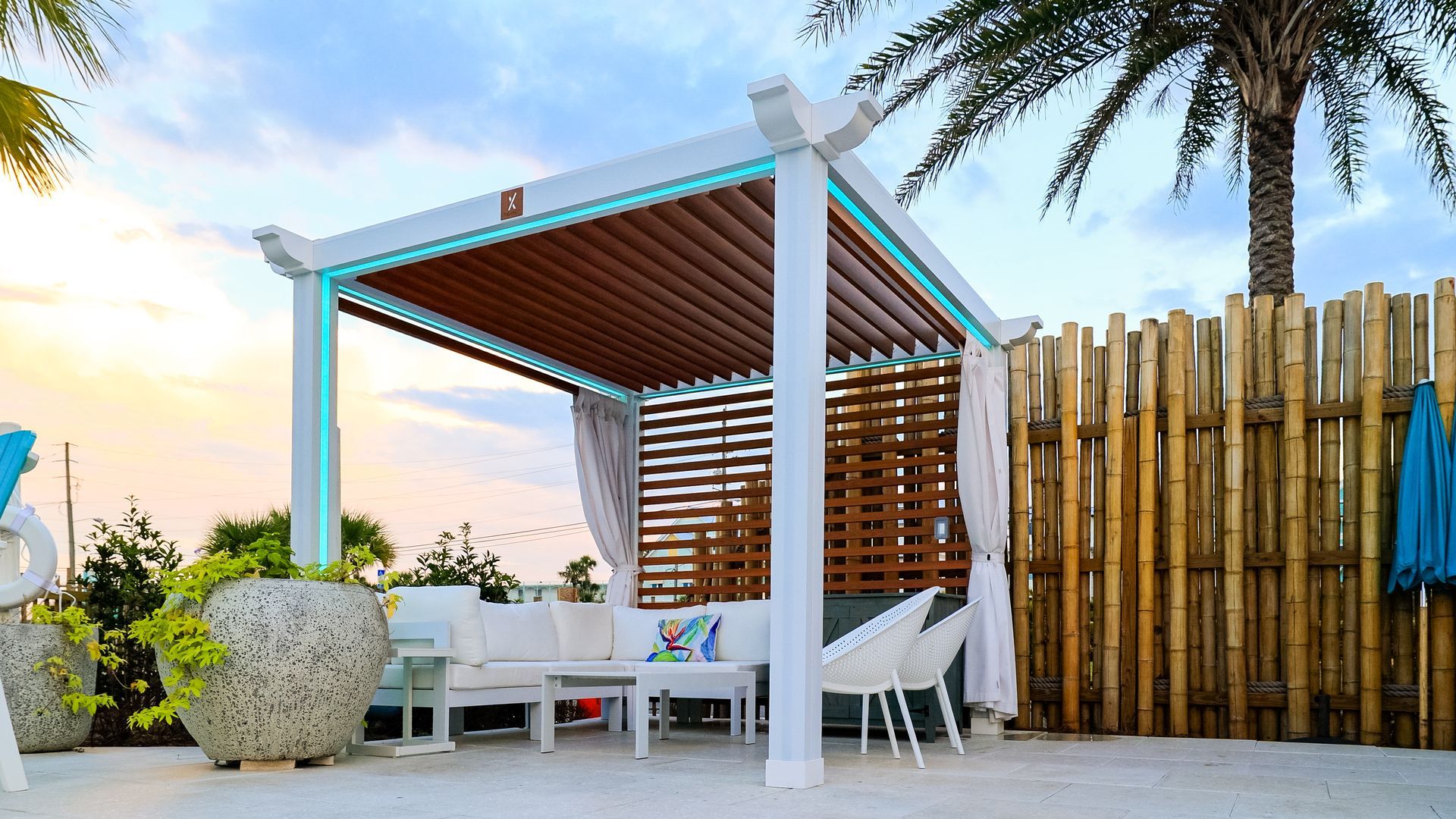 Pergola with white furniture, wooden slats, and curtains on a patio near bamboo fencing and a palm tree.