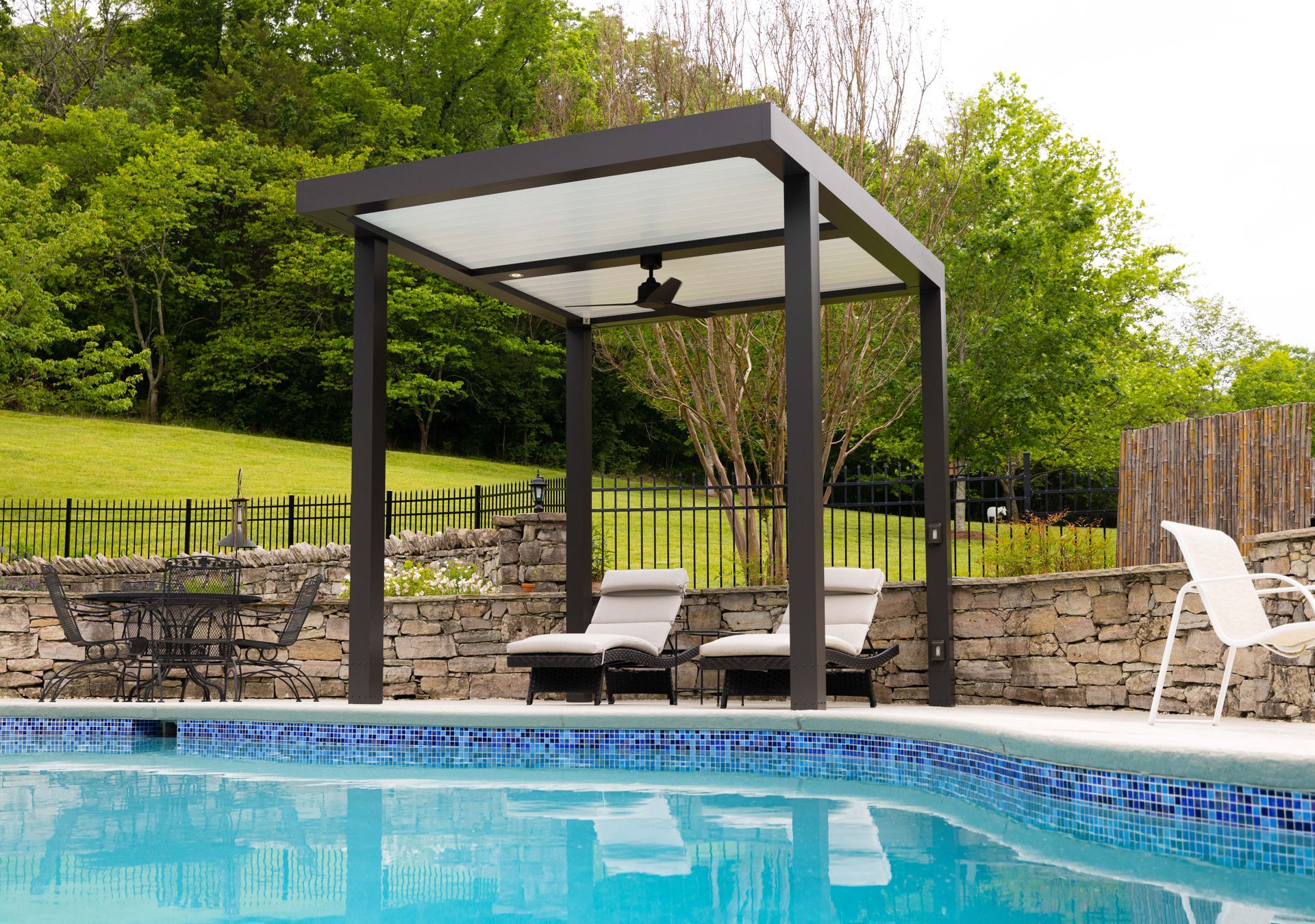 Pergola over pool with two chaise lounges. Trees and stone wall in the background.