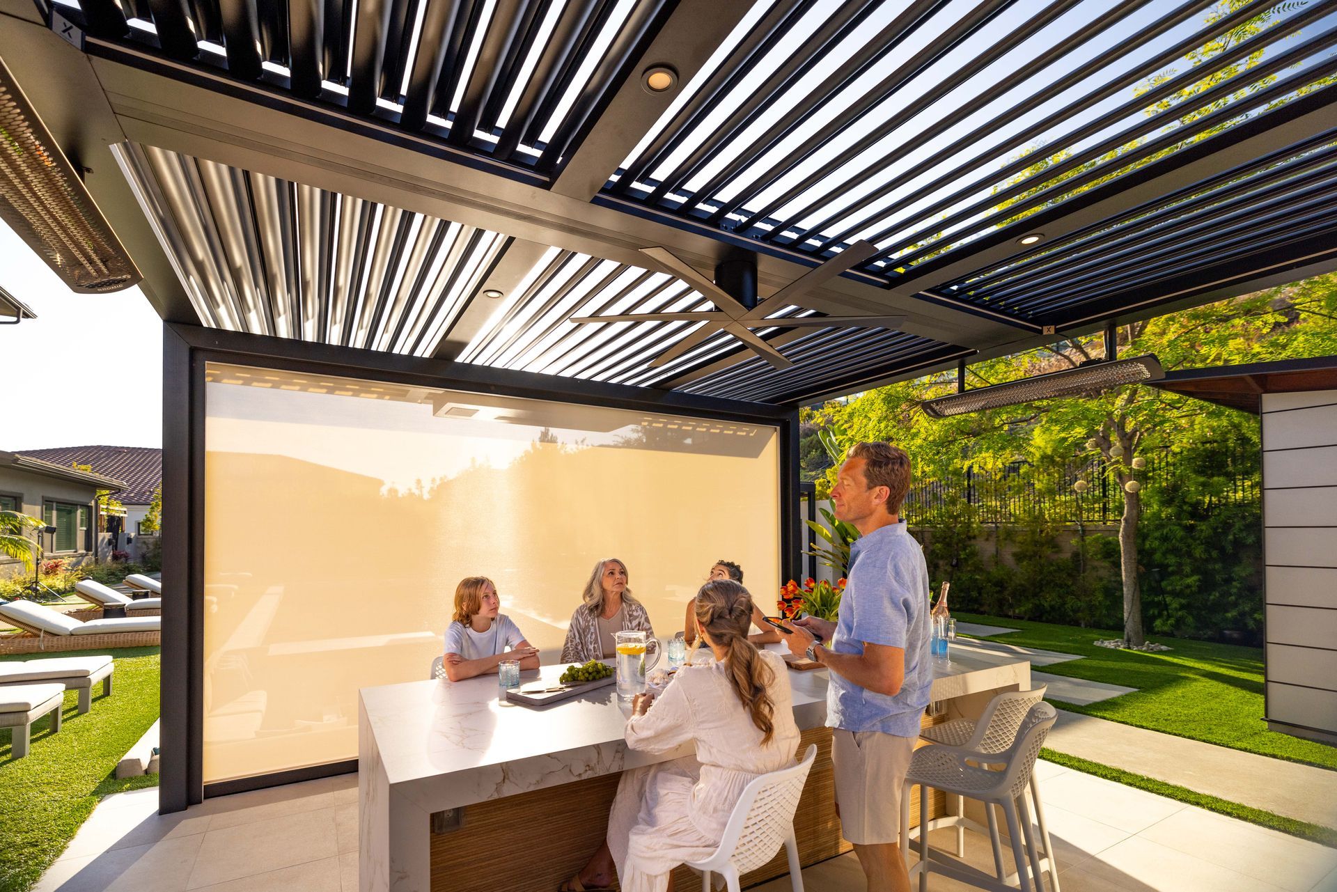 Outdoor patio with a family gathered at a concrete table, partially shaded by an adjustable louvered roof.