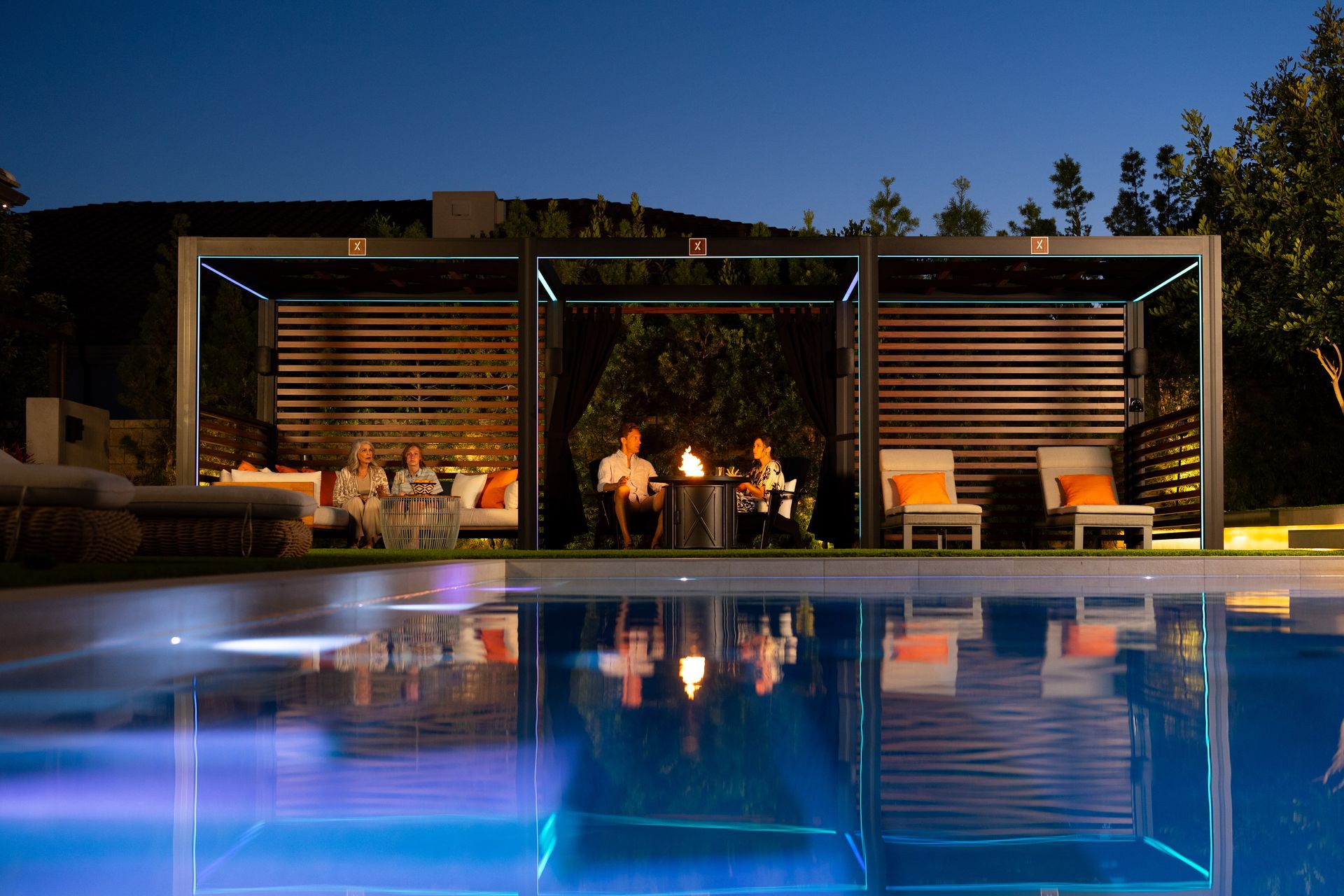 Poolside cabanas at night with people gathered around a fire, reflected in the blue water.