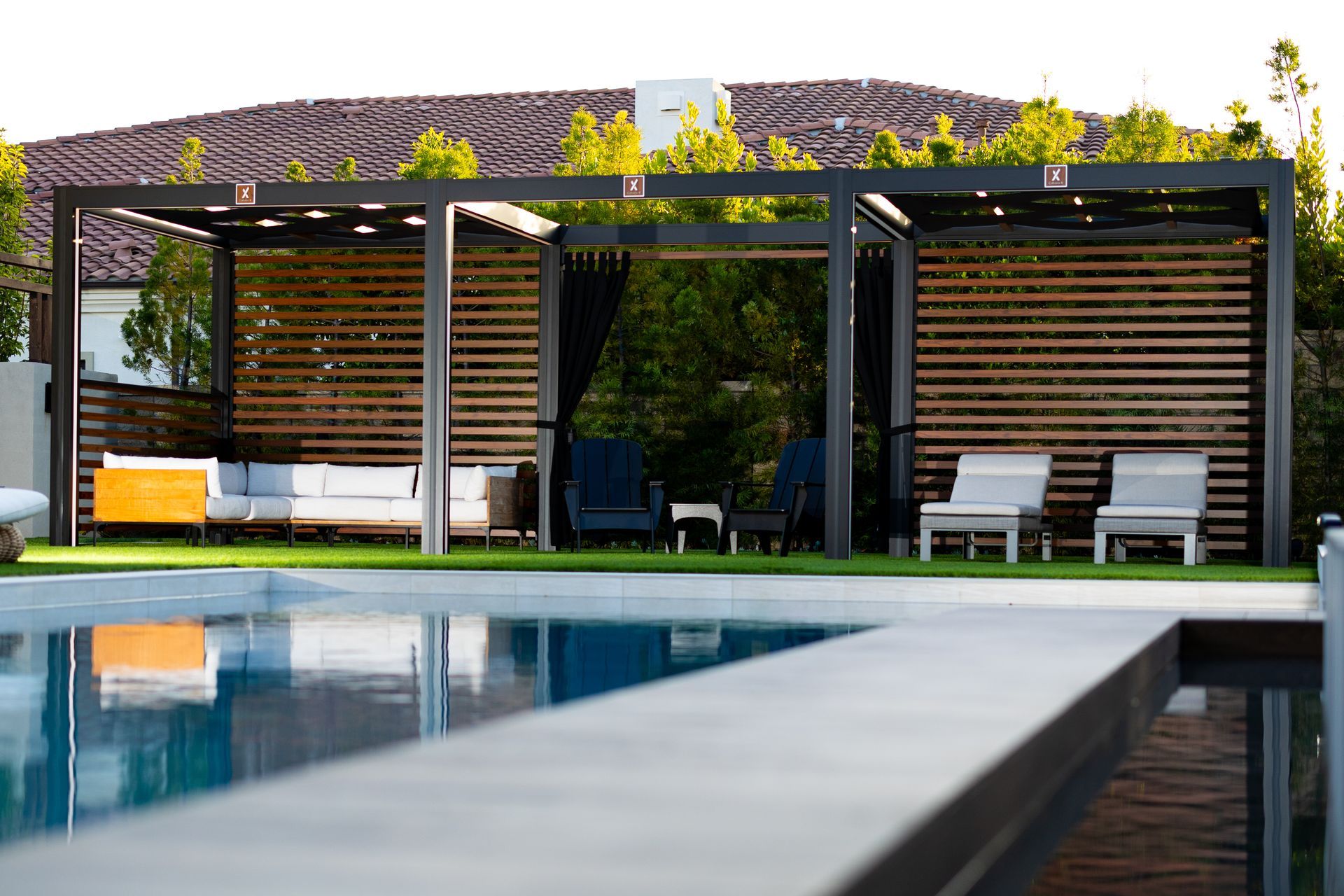 Poolside cabana with seating: couches, chairs, and wooden slatted walls. A pool is in the foreground.