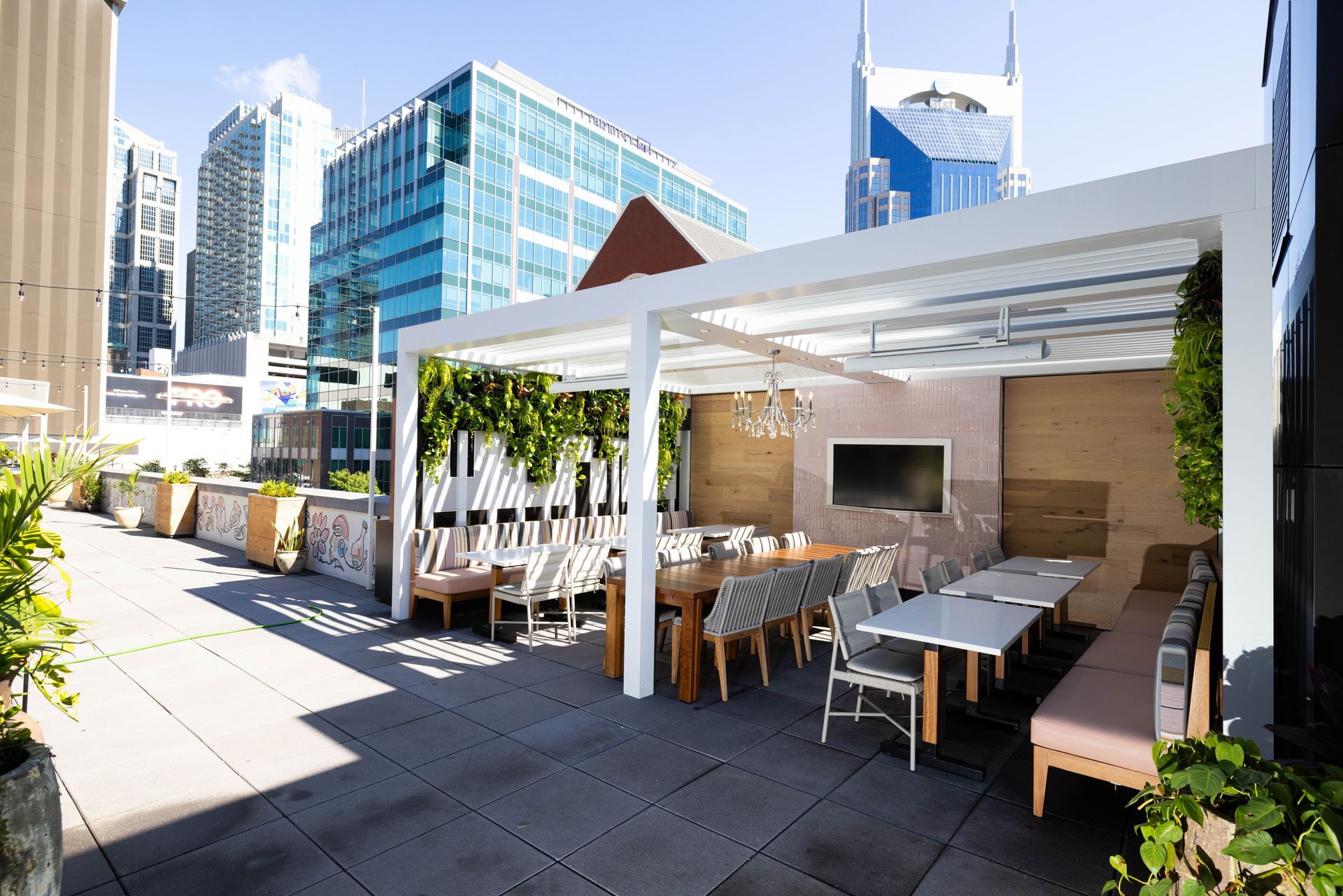 Rooftop patio with seating, tables, and a tv, with city buildings in the background on a sunny day.
