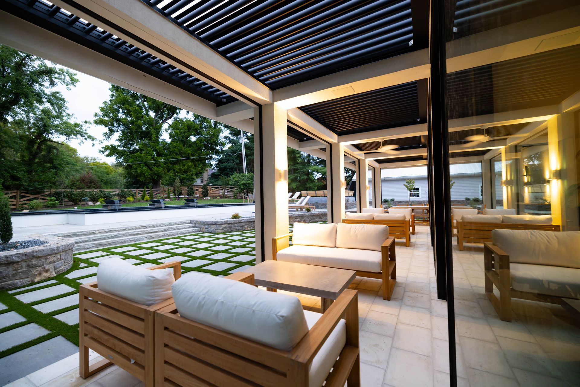 Patio with seating, outdoor view. Wooden furniture, white cushions. Black pergola. Stone tile floor, green lawn.