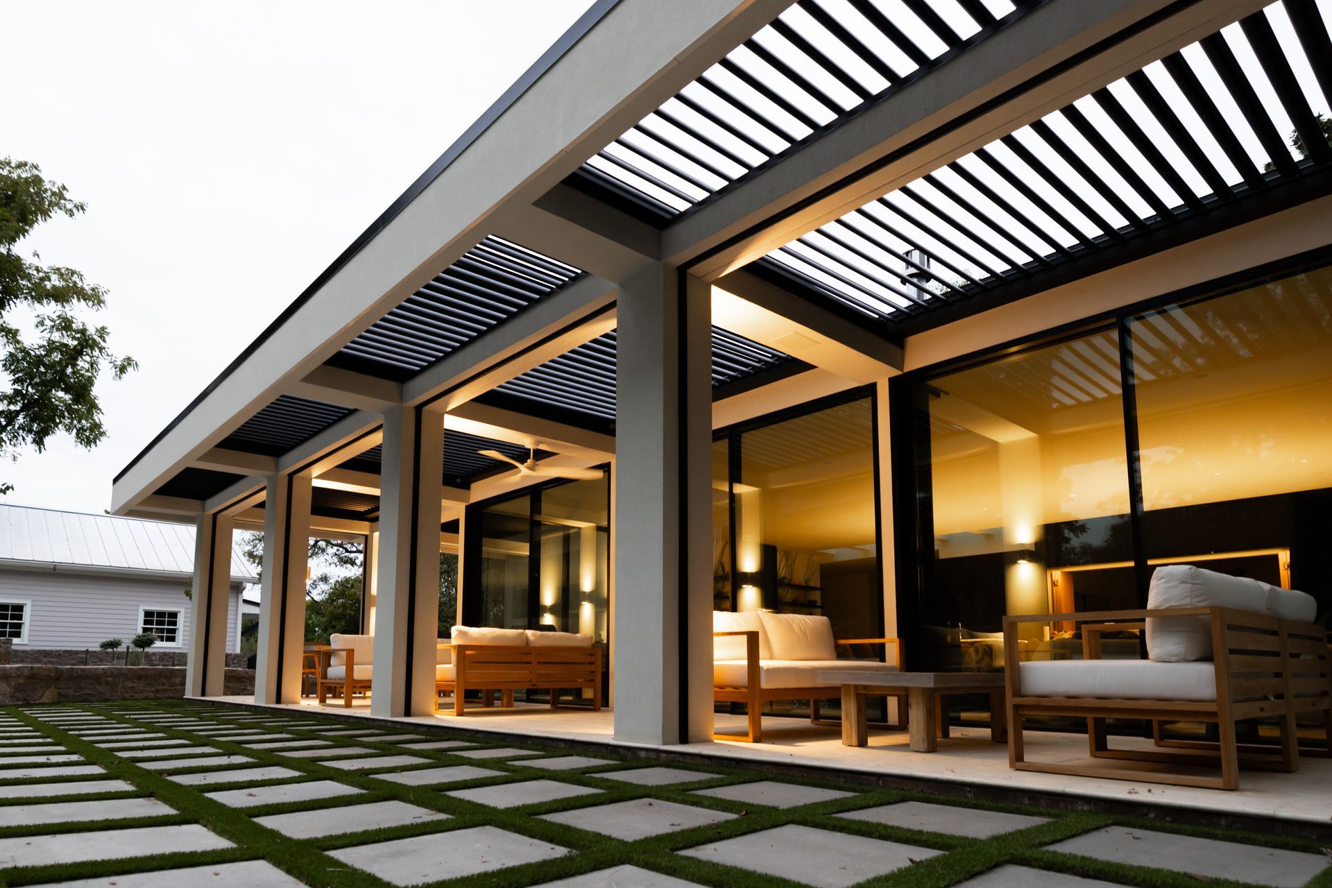 Covered outdoor patio with open beams and seating, concrete pavers with grass, glass doors, and soft interior lighting.