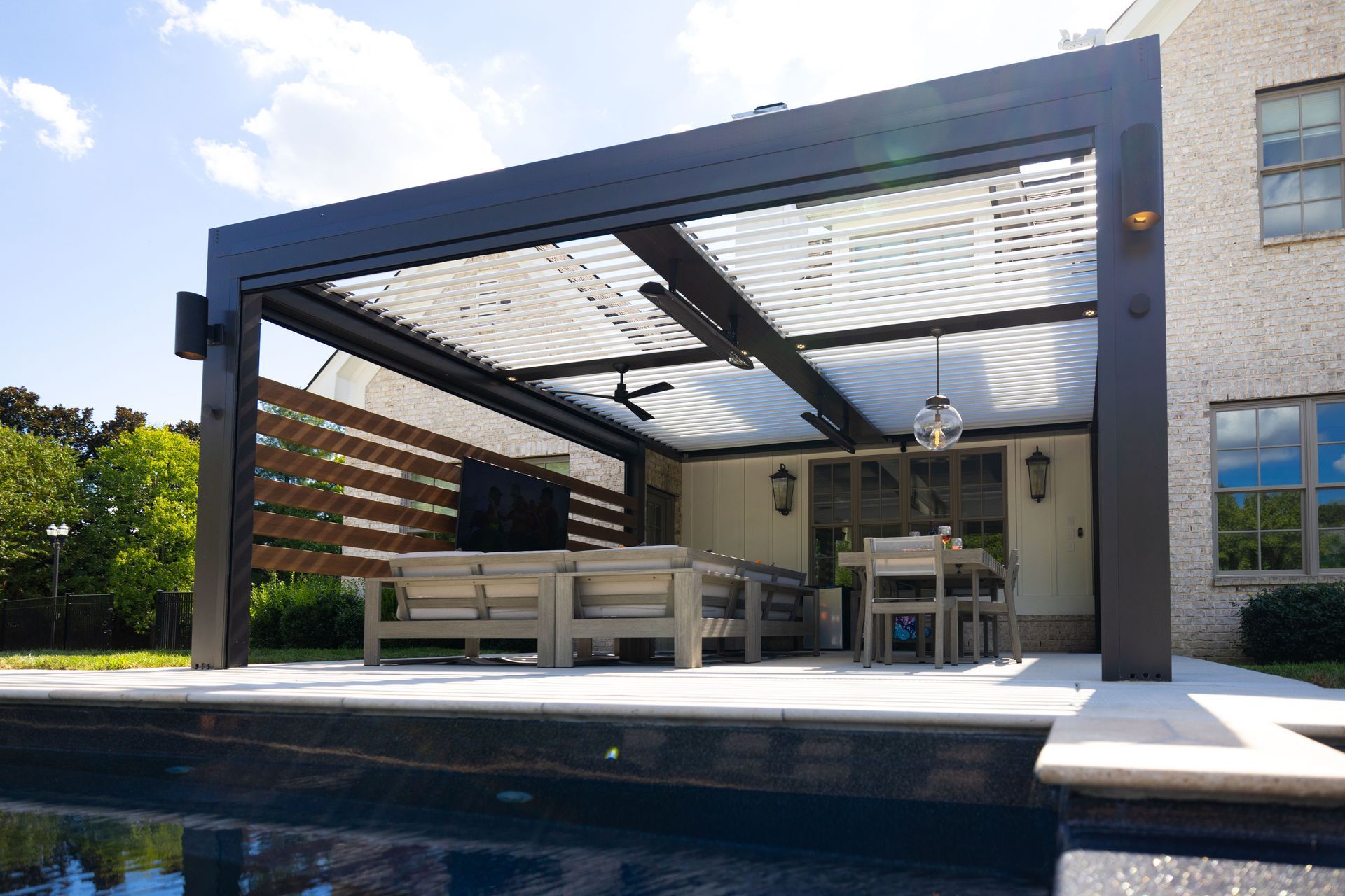 Black pergola over a patio with a pool. Furnished with couches, table, and TV.
