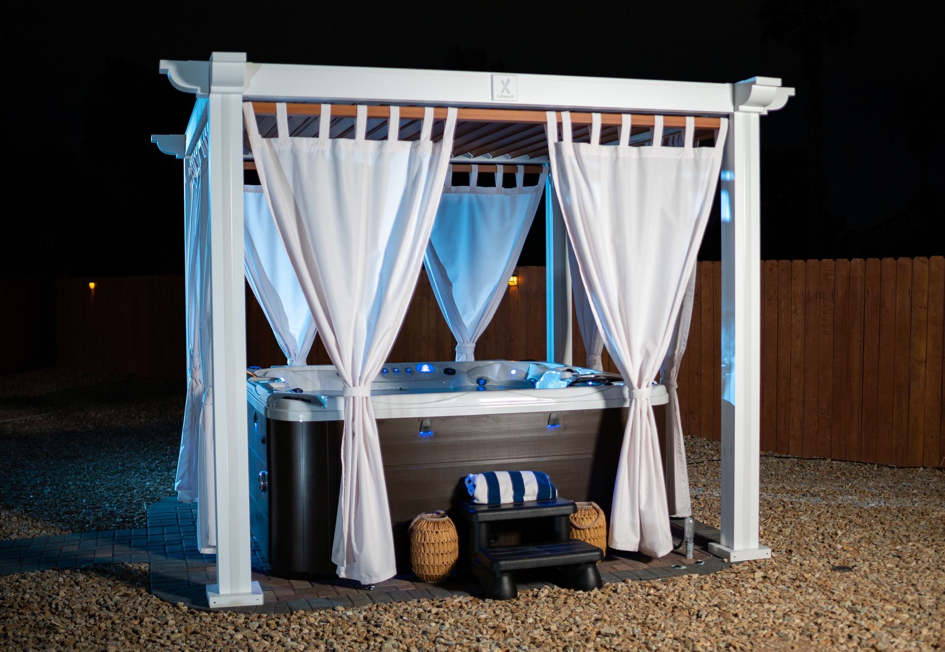 Hot tub in an outdoor wooden gazebo, white curtains, blue lights, dark night setting.