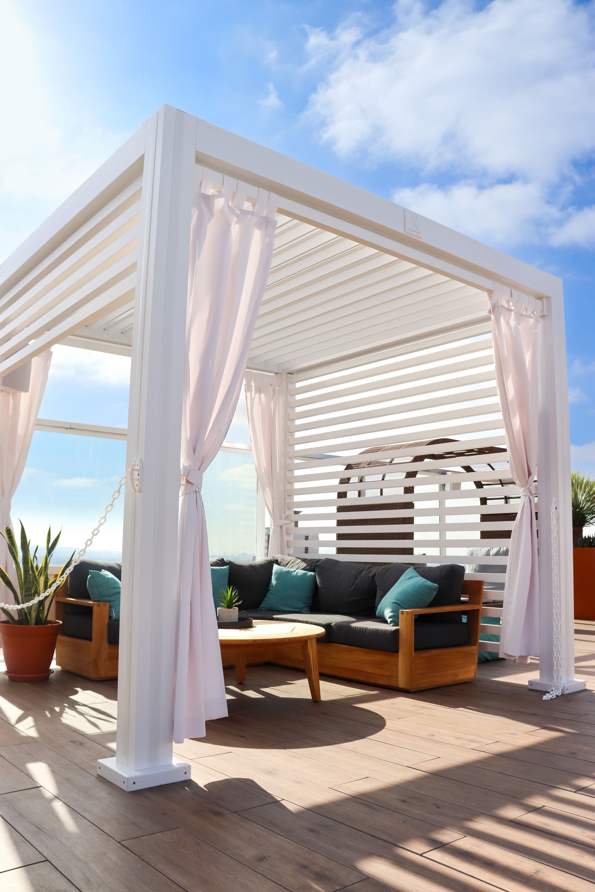 White pergola with curtains, overlooking ocean.  A wooden couch with teal pillows is inside.