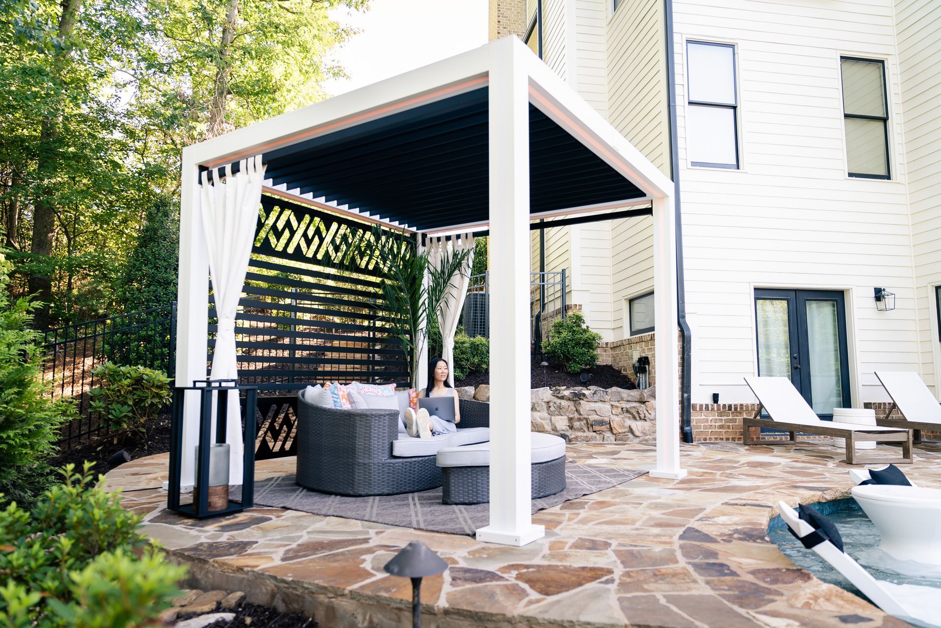 Pergola with seating area next to a pool. A woman sits on a round couch.