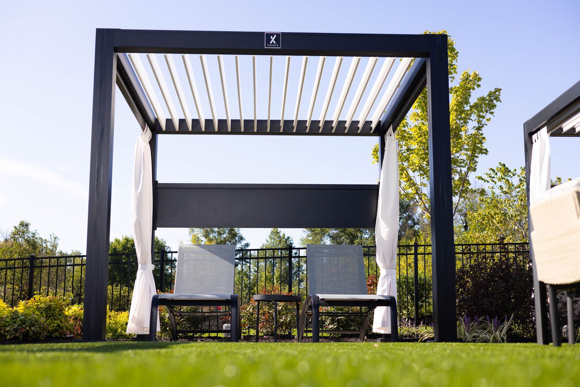 Black pergola with white curtains, two chairs, and small table on green grass.